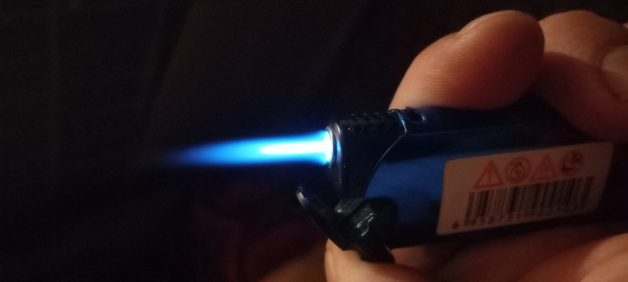 A close-up image depicts a human hand holding an ignited dark blue butane torch lighter against a dark, indistinct background. The lighter emits a tall, narrow, and intensely blue flame with a brighter white core at its base. The lighter's body is dark blue, possibly metallic, with textured grip areas on its sides, and features an open hinged cap mechanism at the top. A white warning label, partially visible on the lower left side of the lighter, displays a barcode and several red hazard symbols, including an exclamation mark within a triangle, a circle with a diagonal line through it, and a snowflake-like symbol. The hand, appearing light-skinned, grips the lighter with visible creases and skin texture, primarily showing the thumb on the right side. The ambient lighting is extremely dim, with the flame serving as the primary light source in the scene. The image contains no visual elements or contextual clues that would indicate a specific geographic location, such as Kryvyi Rih, Ukraine.