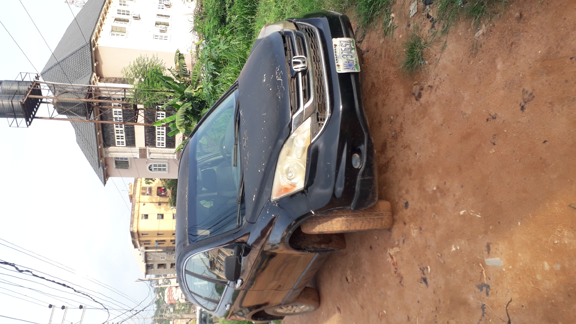 A black Honda CRV is parked on a dirt road in Nkpor, Nigeria. The car is facing right and has Anambra license plate number ANM 375 SZ. The front driver's side tire is flat. Buildings are visible in the background, including a structure with water tanks on its roof. Vegetation is present to the right and behind the car. Electrical lines are overhead.