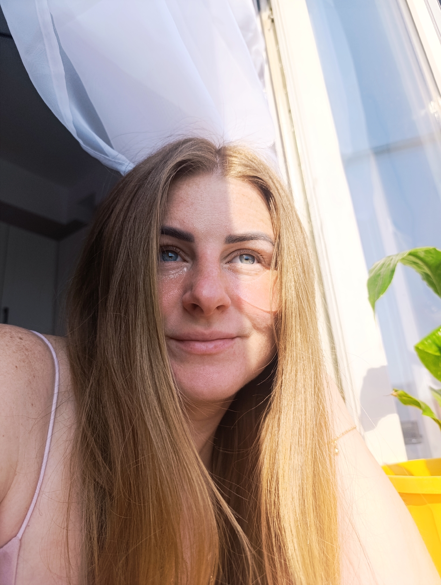 A fair-skinned woman with long blonde hair is centered in a close-up shot, her face slightly wet with what appear to be tears on her cheek. Her blue eyes are looking upward and to the right, and she has a gentle smile. The background shows a window with sheer white curtains on the left and reflective glass panes filled with a blue sky on the right. A green plant is partially visible near the window, and a yellow object is present in the lower right corner. A thin-strapped top suggests she is indoors in a casual setting, possibly in the city of Unknown, Unknown.
