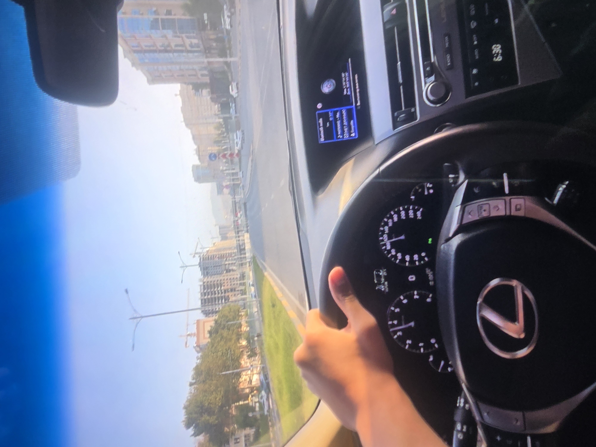 The image, taken from the perspective of a driver, shows a first-person view of driving a Lexus. The driver's hand is visible on the steering wheel, which prominently features the Lexus logo. The dashboard displays the car's instruments, including a speedometer and tachometer, with the needle of the speedometer indicating a speed of approximately 50-60 km/h. The car's infotainment system is also visible, displaying "Bluetooth audio" and other information. Outside the car, the scene is an urban environment with buildings and trees lining a road. Construction is underway on some of the buildings. The sky is clear and blue, suggesting it is daytime. The lighting indicates it might be late afternoon due to the warm tones on the driver's hand.