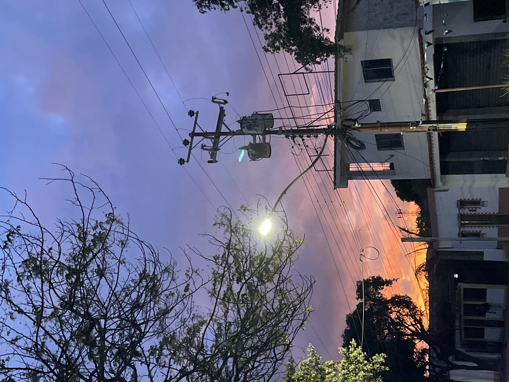 This is a vertical, medium shot image taken at dusk in an urban environment, possibly in the city of Unknown, Unknown. The sky displays a gradient of colors from deep purple at the top to vibrant orange and pink near the horizon, suggesting a sunset or sunrise. A large, bare tree with thin branches dominates the left side of the frame, partially obscuring the sky. A street lamp with a bright light is visible on the right side of the tree. A utility pole with electrical transformers and numerous wires stands prominently in the center of the image, running vertically through the frame. In the foreground, a white building with a dark awning and closed shutters is visible. To the left of this building, another dark building with windows and what appear to be closed storefronts can be seen. The overall scene depicts a residential or commercial area at twilight, with a focus on the interplay of natural light, urban infrastructure, and residential structures. There are no people visible in the image.