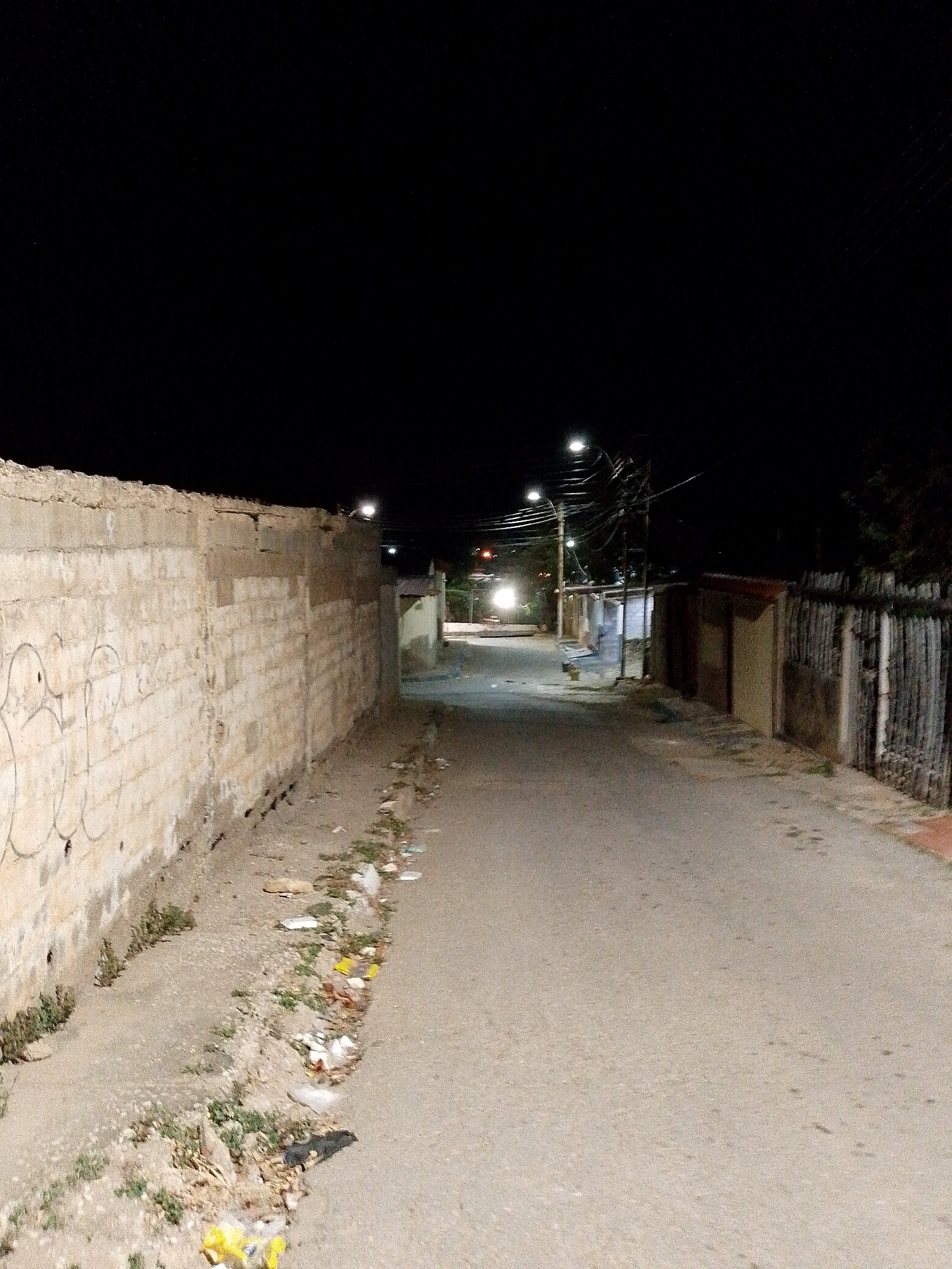 The image depicts an empty street at night in what appears to be Juan Griego, Venezuela. The street is flanked on one side by a high, weathered concrete block wall covered with graffiti. On the other side is a mix of buildings and fences. The street surface is paved, but shows signs of wear. Trash and debris are visible along the edges of the street. Streetlights illuminate the scene, casting long shadows down the road. Overhead, electrical wires are strung between poles. A light source further down the street suggests a vehicle or another source of illumination at the end of the road. No people are visible.