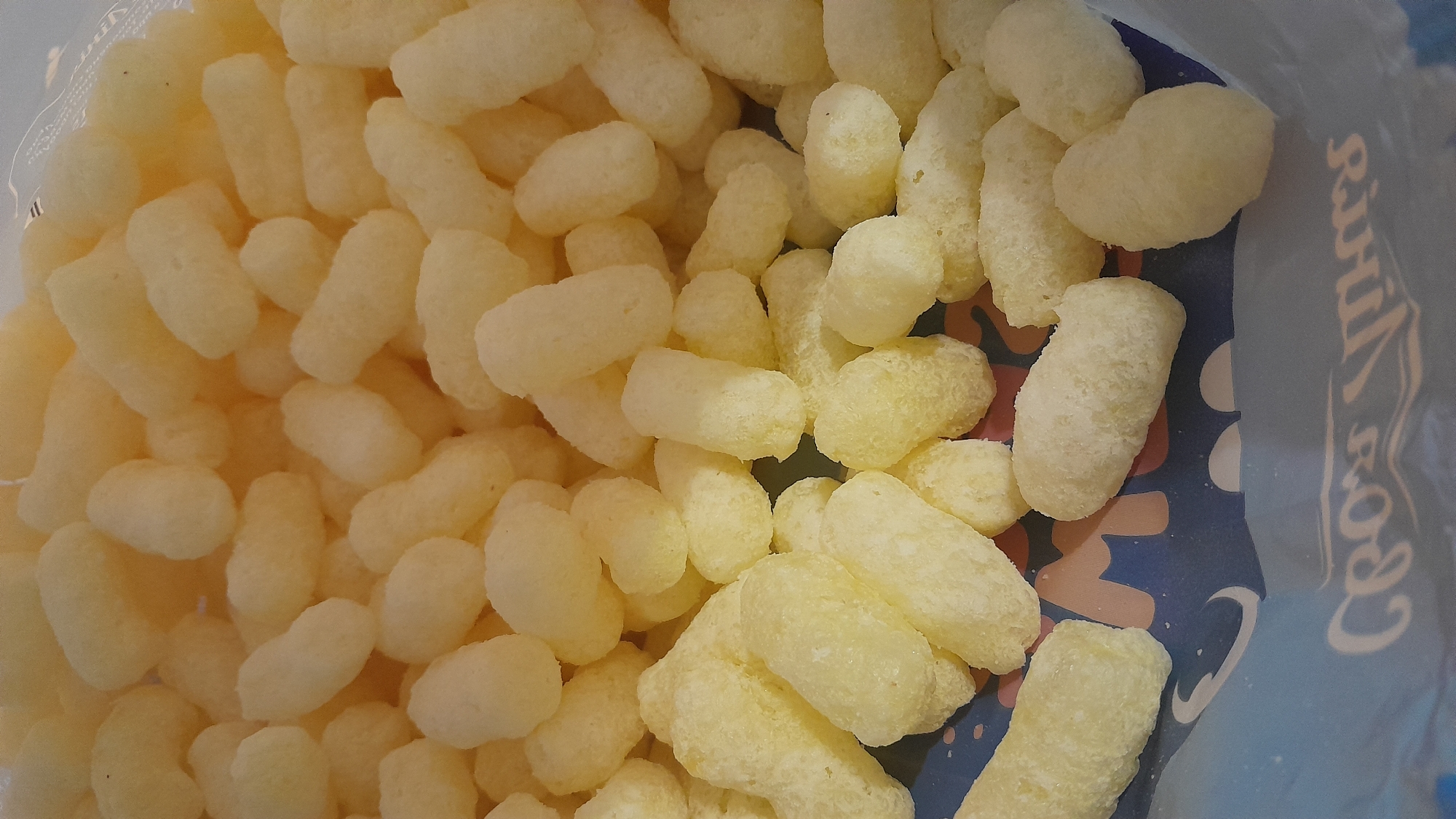 A close-up, overhead shot captures a pile of light yellow, puffy, cylindrical snack pieces. The snacks, resembling corn puffs or cheese puffs, are densely packed together, filling most of the frame. The texture of the snacks appears airy and slightly crumbly. The background shows a portion of blue packaging with some text and graphic elements visible, partially obscured by the snacks. The text appears to be white, stylized lettering, possibly part of a brand name or slogan. The lighting is bright, highlighting the texture and color of the snacks. The scene is devoid of any people or recognizable city elements, making it impossible to determine a location within a city.