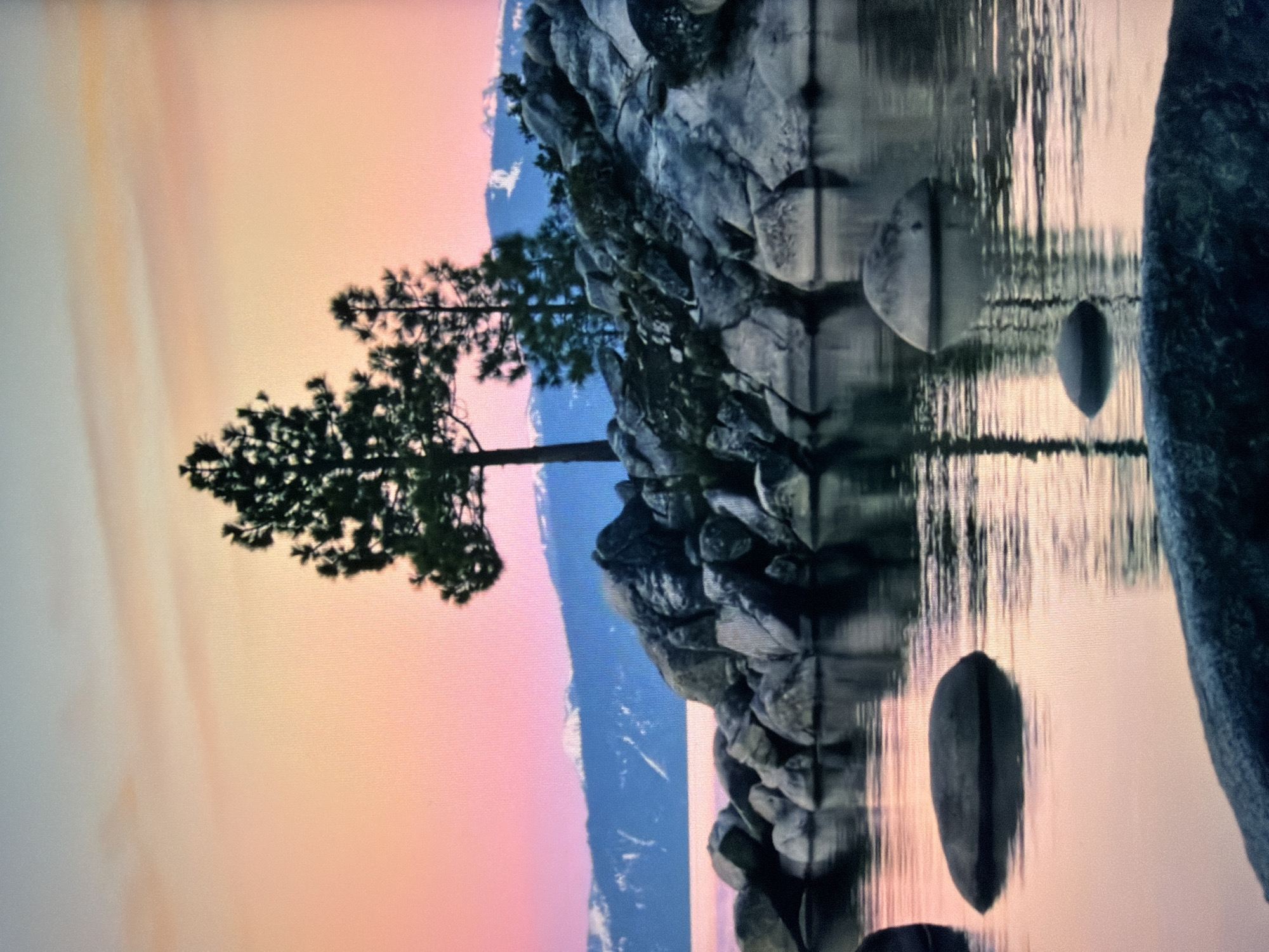 This is a photograph of a serene natural landscape at sunset. The sky displays a gradient of soft colors, transitioning from a pale blue at the top to shades of pink and orange towards the horizon. In the foreground, a body of calm water reflects the sky and the surrounding elements.
Dominating the middle ground is a rocky shoreline. Large, grey boulders of various sizes are piled along the edge of the water. Several evergreen trees, silhouetted against the sky, grow from the rocks. The largest tree stands tall and prominent, with a second, smaller tree to its right.
In the background, across the water, is a mountain range with snow-capped peaks. The water's surface is smooth, with subtle ripples creating distorted reflections of the trees and rocks. The overall atmosphere is tranquil and picturesque. There are no people or any indications of human activity in this scene. This image does not depict any location in the city of Kyiv, Ukraine; it is a natural landscape.