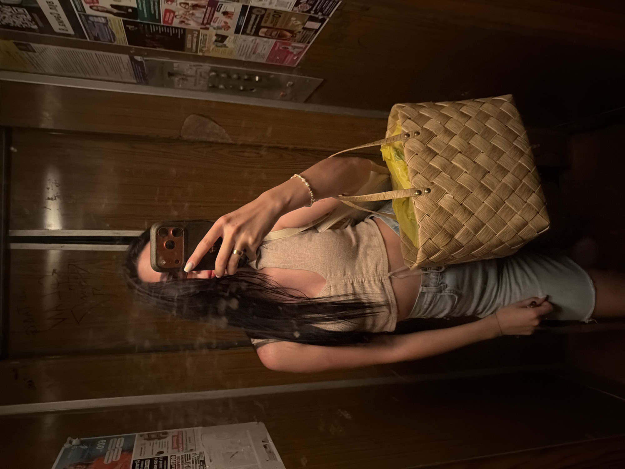 In an elevator, a person with long dark hair takes a mirror selfie. They are wearing a light beige, ribbed crop top with a drawstring detail at the front and a light blue denim skirt. A woven straw basket with yellow contents is held in their right arm, and a bracelet adorns their left wrist. The elevator's walls are a dark wood paneling, with some graffiti visible on the left wall. A bulletin board with various papers and advertisements is mounted on the right wall, near the elevator control panel.