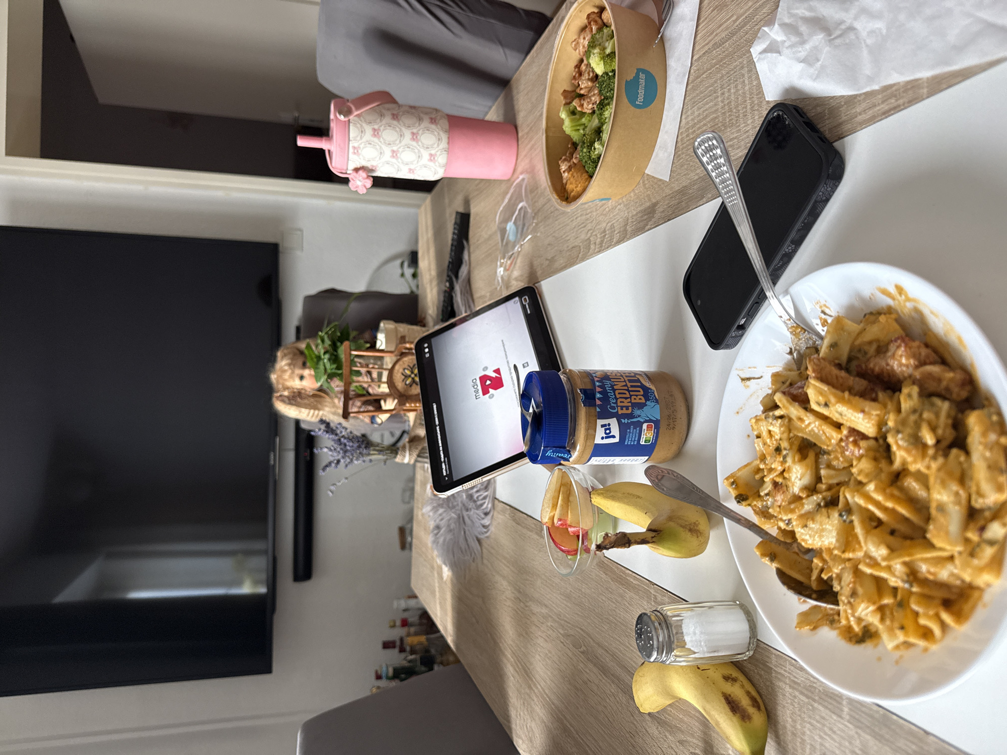 The image depicts a dining table set for a meal, likely in a home in Hanau, Germany. In the foreground, a plate of pasta is visible, accompanied by a spoon. To the left, there are two bananas, a salt shaker, and a bowl of sliced apples. A jar of peanut butter stands next to the pasta. In the mid-ground, a tablet displays a YouTube logo. A pink sippy cup with a floral pattern sits to the right. Further back, a doll is seated on a small chair in front of a large television screen. The environment appears to be a living or dining area, with a wooden table and chairs. The lighting suggests it is daytime.