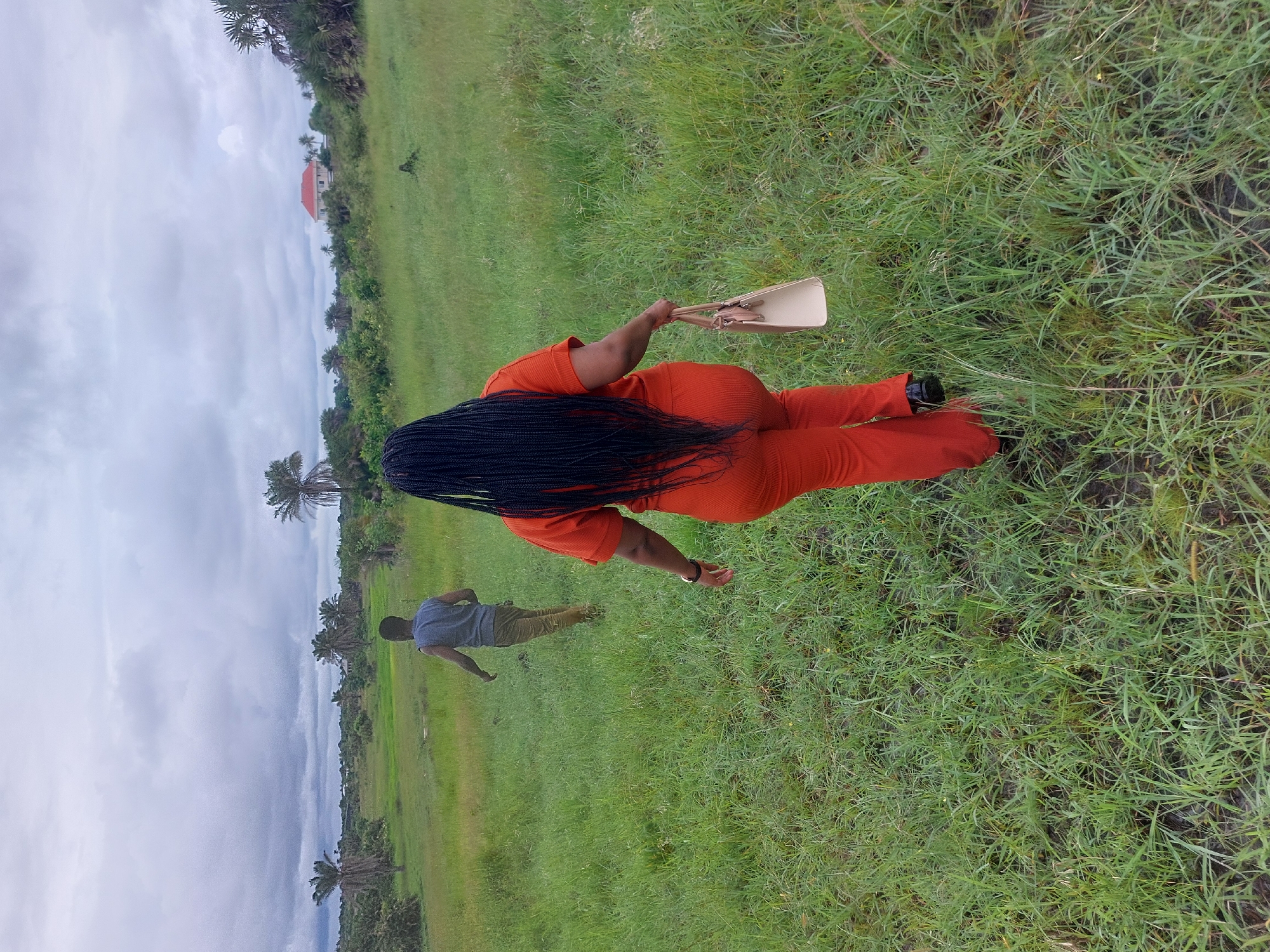 The image captures a scene in what appears to be a grassy field. A woman is prominently featured from the back, wearing an orange outfit and holding a beige purse. Her long black braided hair extends down her back. A man wearing a gray shirt and khaki pants walks in the background. The environment consists of green grass and vegetation. In the background, there are trees and a building with a red roof. The cloudy sky suggests a rural or semi-rural setting. Without further data points, it is not possible to determine if this scene is in Dar es-Salaam, Tanzania.