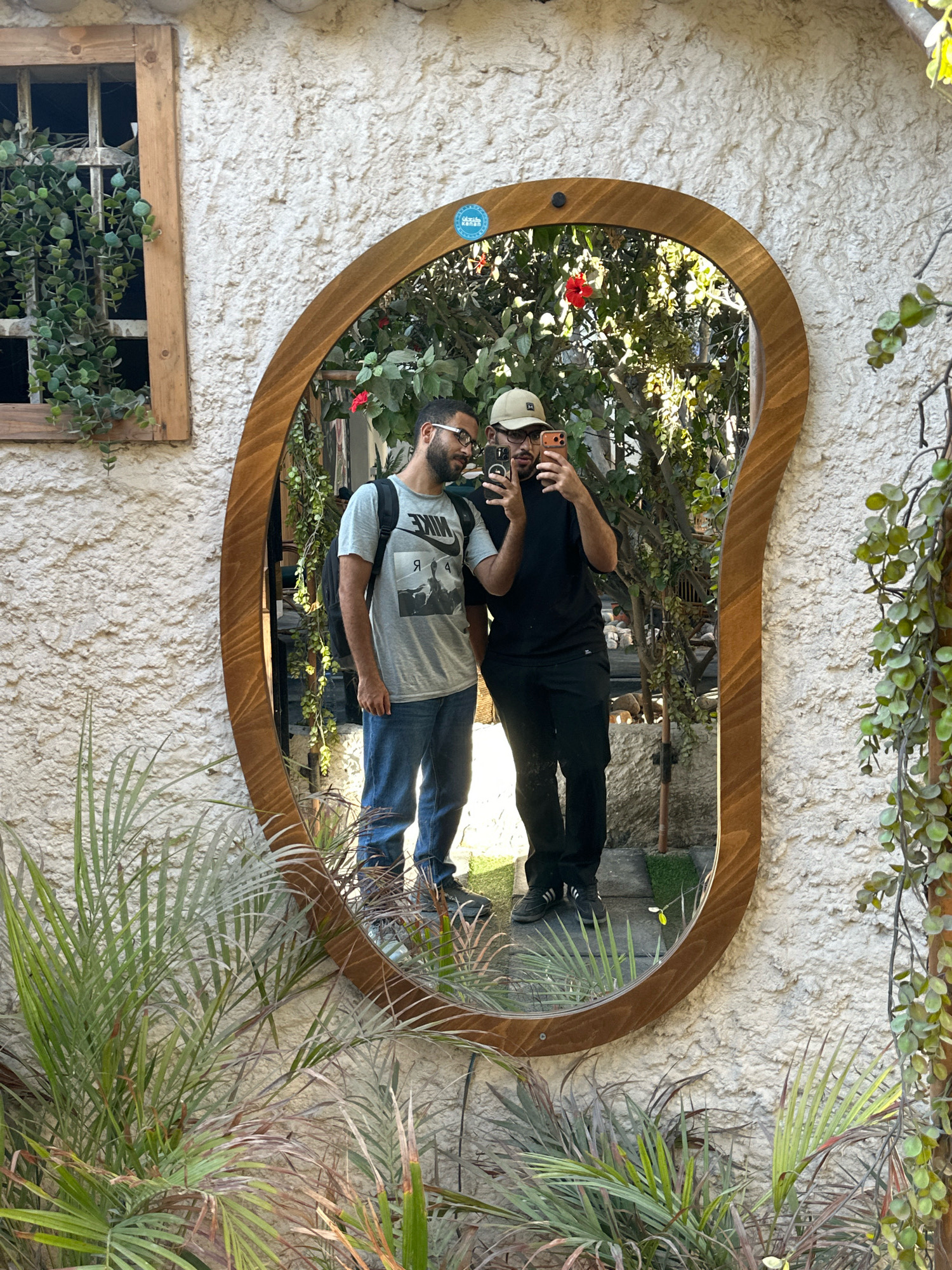 In the city of Gaza, Palestinian Territories, a person is taking a mirror selfie in front of a textured, off-white wall. The mirror, framed with natural wood and irregularly shaped, reflects two individuals. One person, on the left, wears a gray t-shirt with a graphic print, blue jeans, glasses, and a backpack. The other person, on the right, is dressed in a black t-shirt, black pants, and a beige baseball cap, holding a smartphone. Behind them, lush greenery and a red flower are visible through the mirror's reflection, suggesting an outdoor setting. In the foreground and to the sides of the mirror, various potted plants with long, green leaves are present. To the left of the mirror, a wooden-framed window with bars contains more hanging greenery.