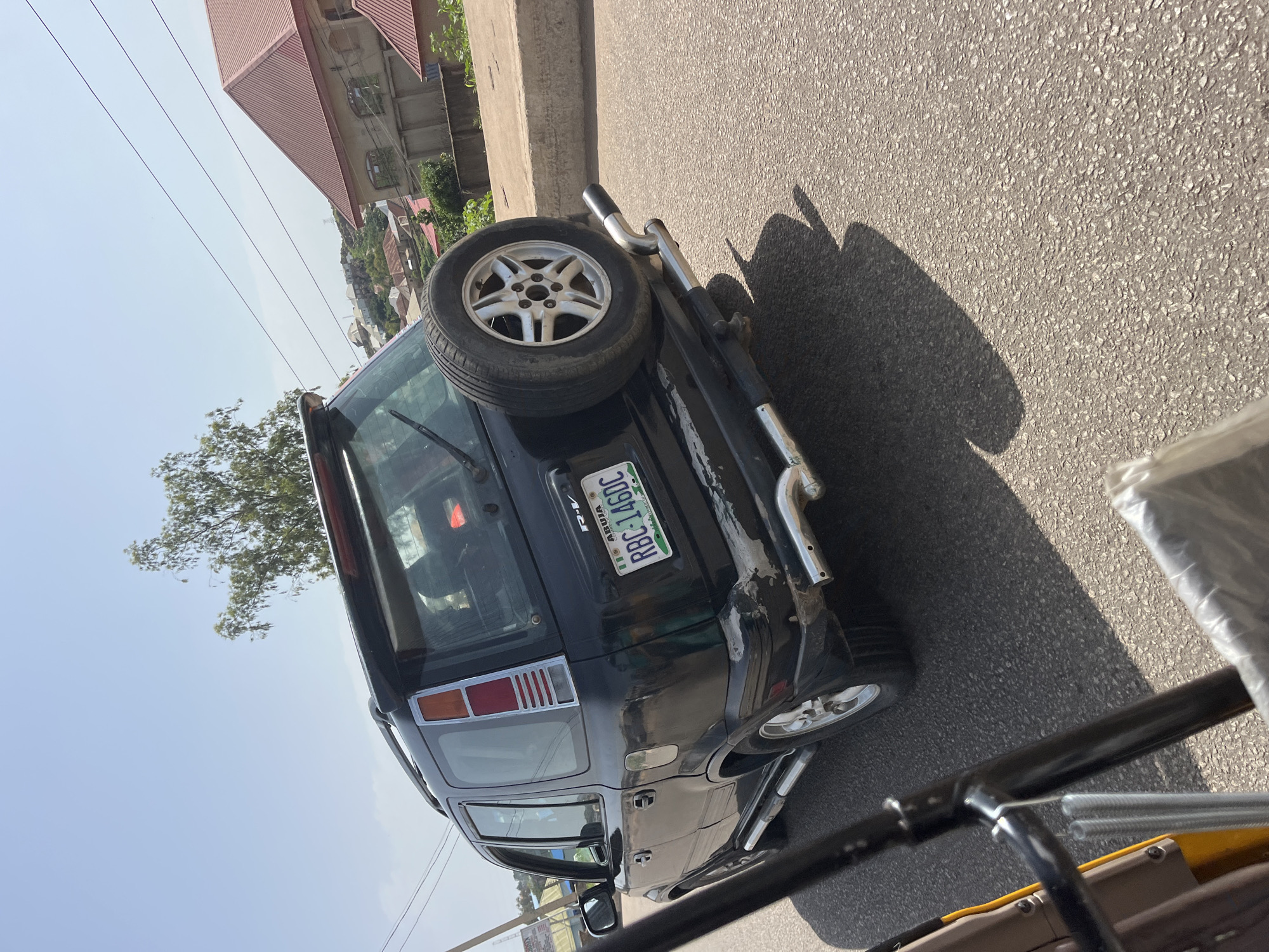A black Honda CR-V is parked on the side of a road. The rear of the vehicle is visible, showing a spare tire mounted on the tailgate, a rear bumper with chrome accents, and a license plate that reads "RBC-146DC". The vehicle's paint is slightly worn, particularly around the rear bumper. The license plate indicates it is registered in Abuja. The scene appears to be a sunny day, with a clear sky and shadows cast on the asphalt road. In the background, there are residential buildings and trees. The image is taken from a low angle, possibly from another vehicle.