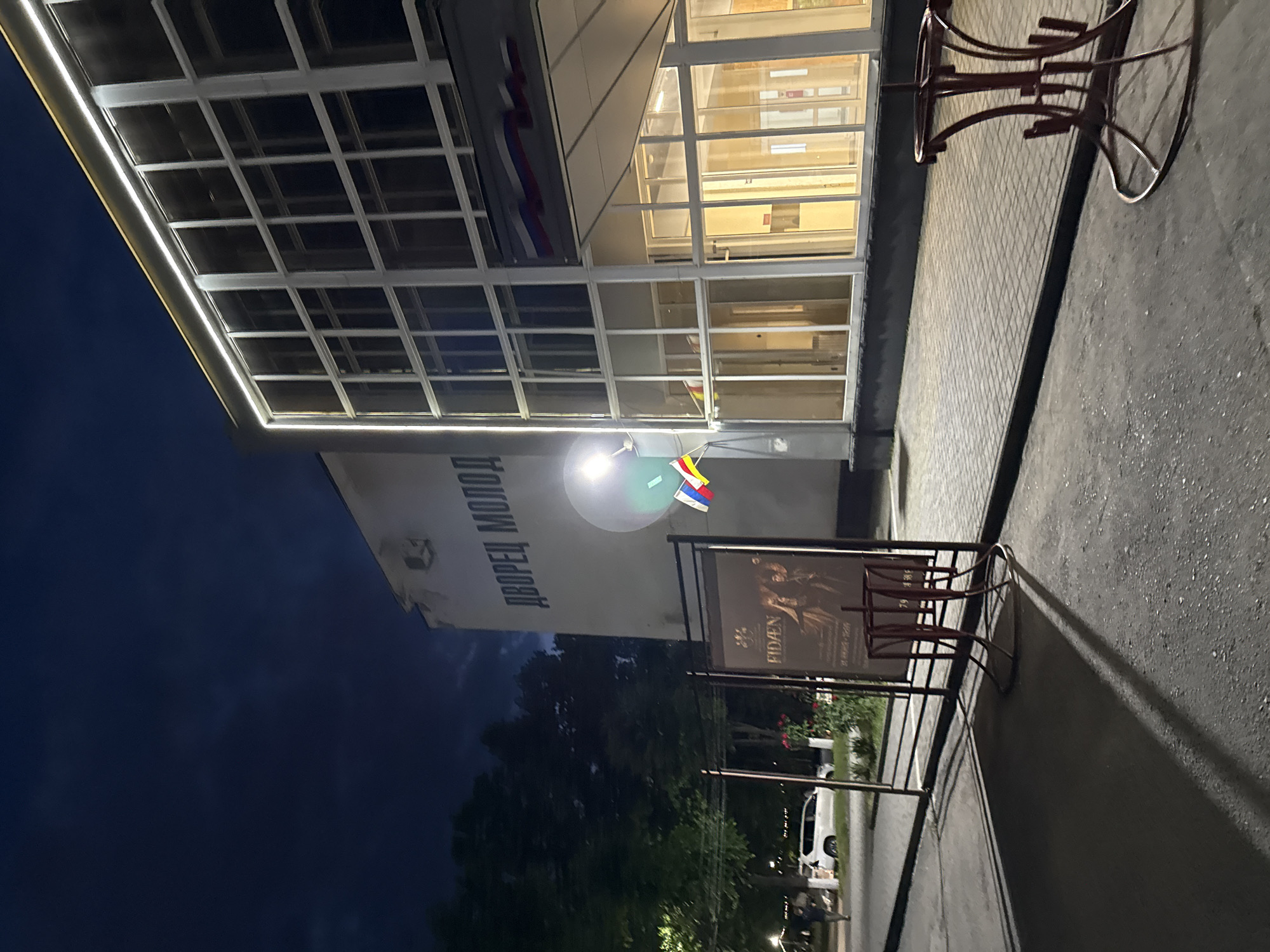 The image shows the entrance to the Palace of Youth in Vladikavkaz, Russia, at night. The building has large windows and is illuminated from within. A sign with Russian text is visible on the facade. In front of the building, there are two posters advertising an event. Flags are also displayed. The scene is lit by artificial lights and the sky is dark, indicating it is nighttime. The ground is paved with bricks and asphalt. A white car is parked nearby, and a few people are visible in the background. The atmosphere appears calm and quiet. The poster text is partially visible, with "RIDEN" being clearly legible. The building's architecture suggests a modern design.