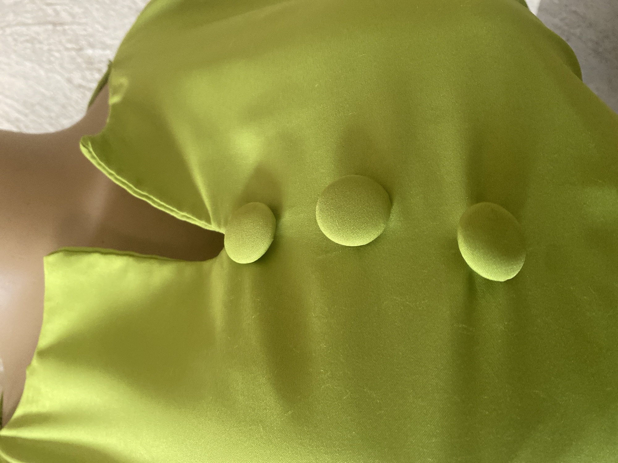 The image shows a close-up of a bright lime green garment with three fabric-covered buttons. The garment appears to be made of a satin-like material, as evidenced by its sheen and the way it drapes. The buttons are round and uniformly spaced. The garment is laid out, possibly on a flat surface or a mannequin, with the texture of a white, possibly terrycloth, fabric visible in the upper left corner. The lighting suggests it's daytime, but the specific time of day or weather conditions are not discernible. No people are visible, and there are no discernible location cues beyond the garment itself. The image is purely focused on the texture and details of the clothing item. There is no visible text in the image.