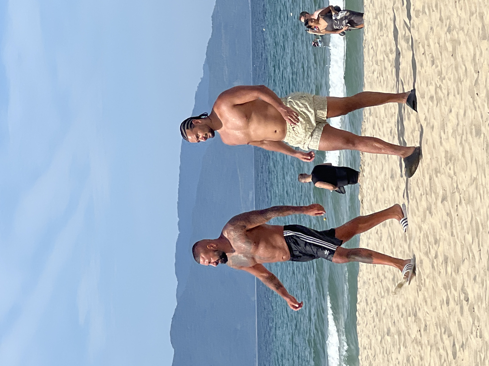 Two men are walking on a sandy beach with the ocean and a mountain range in the background. The man on the left is wearing black Adidas shorts with white stripes and white Adidas flip-flops. He has tattoos on his arms and chest. The man on the right is wearing patterned light-colored shorts and dark slides. He has his hair braided into cornrows. There are other people in the background, some in the water and some on the beach. The sky is blue with some wispy clouds. The scene appears to be in Đà Nẵng, Vietnam, given the context provided, likely near the coast with views of the Sơn Trà Peninsula mountains.