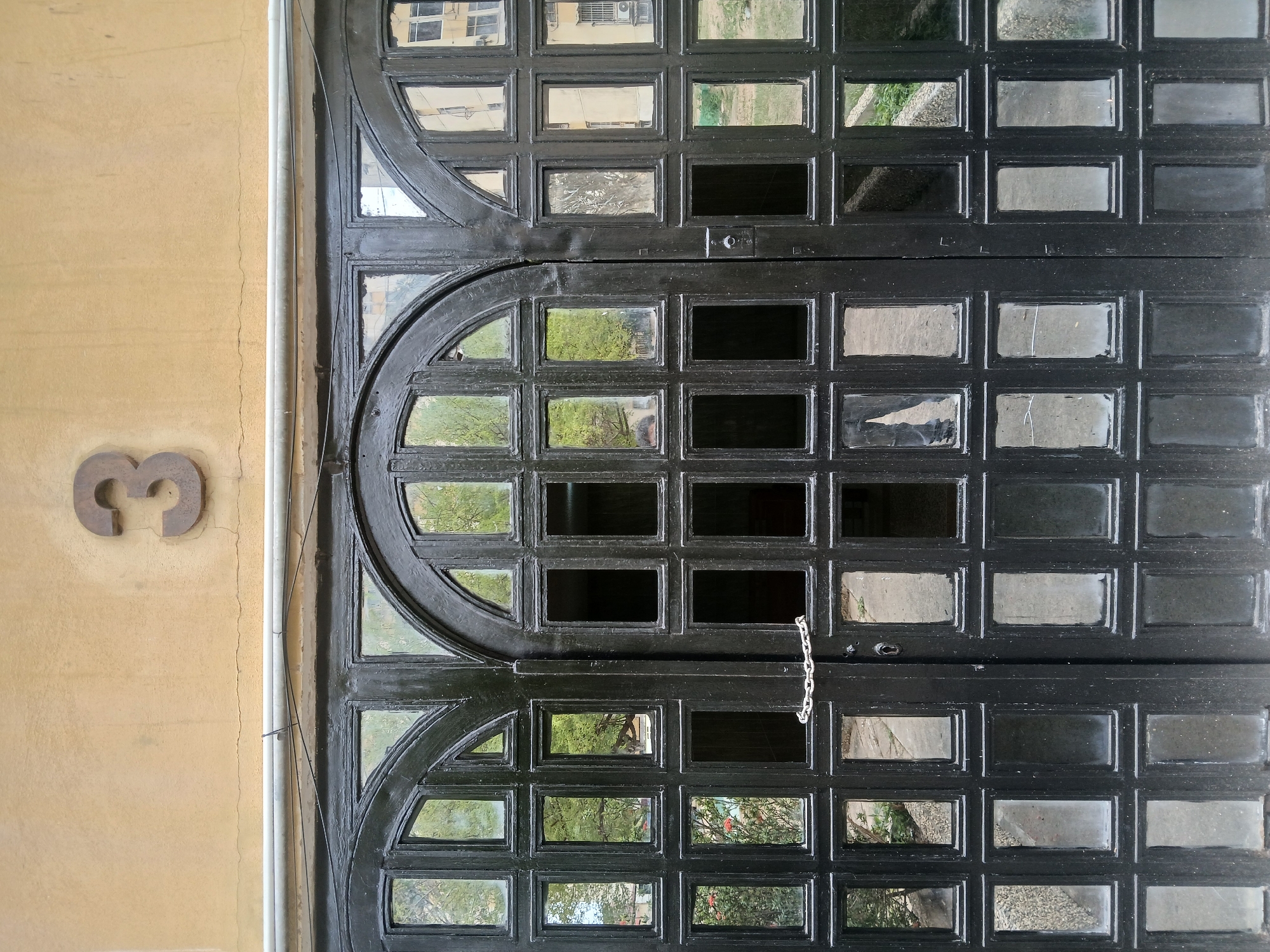 The image depicts a close-up view of a large, black, ornate door, likely an entrance to a building. The door is divided into rectangular and curved sections, each containing glass panes. Some panes appear to be clear, reflecting the exterior environment, while others appear dark or obscured. A metal chain hangs loosely, presumably serving as a security measure or closure mechanism. To the left of the door, affixed to a textured, tan-colored wall, is a metal number "3". A white pipe runs vertically alongside the wall and door frame. The location is Ciudad Guayana, Venezuela. There are no visible people or explicit interactions in the frame. The environment reflected in the door's glass panes suggests an outdoor setting with trees and building structures.