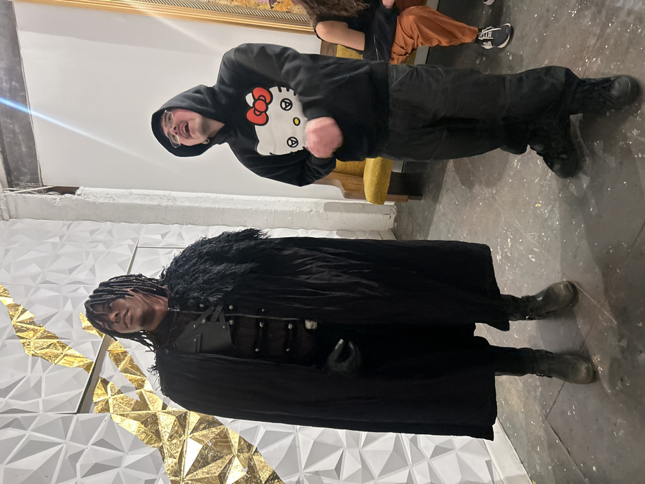 Two individuals pose indoors, seemingly at an event or gathering. One wears a black "Hello Kitty" hoodie, black pants, and dark shoes. The other person is dressed in a long, dark coat with a fur collar and what appears to be a costume-style outfit underneath. A patterned wall serves as a backdrop, with geometric shapes and gold accents visible behind the person in the coat. Part of a gold-framed painting is at the top of the shot. The lighting is bright, and the floor appears to be concrete.