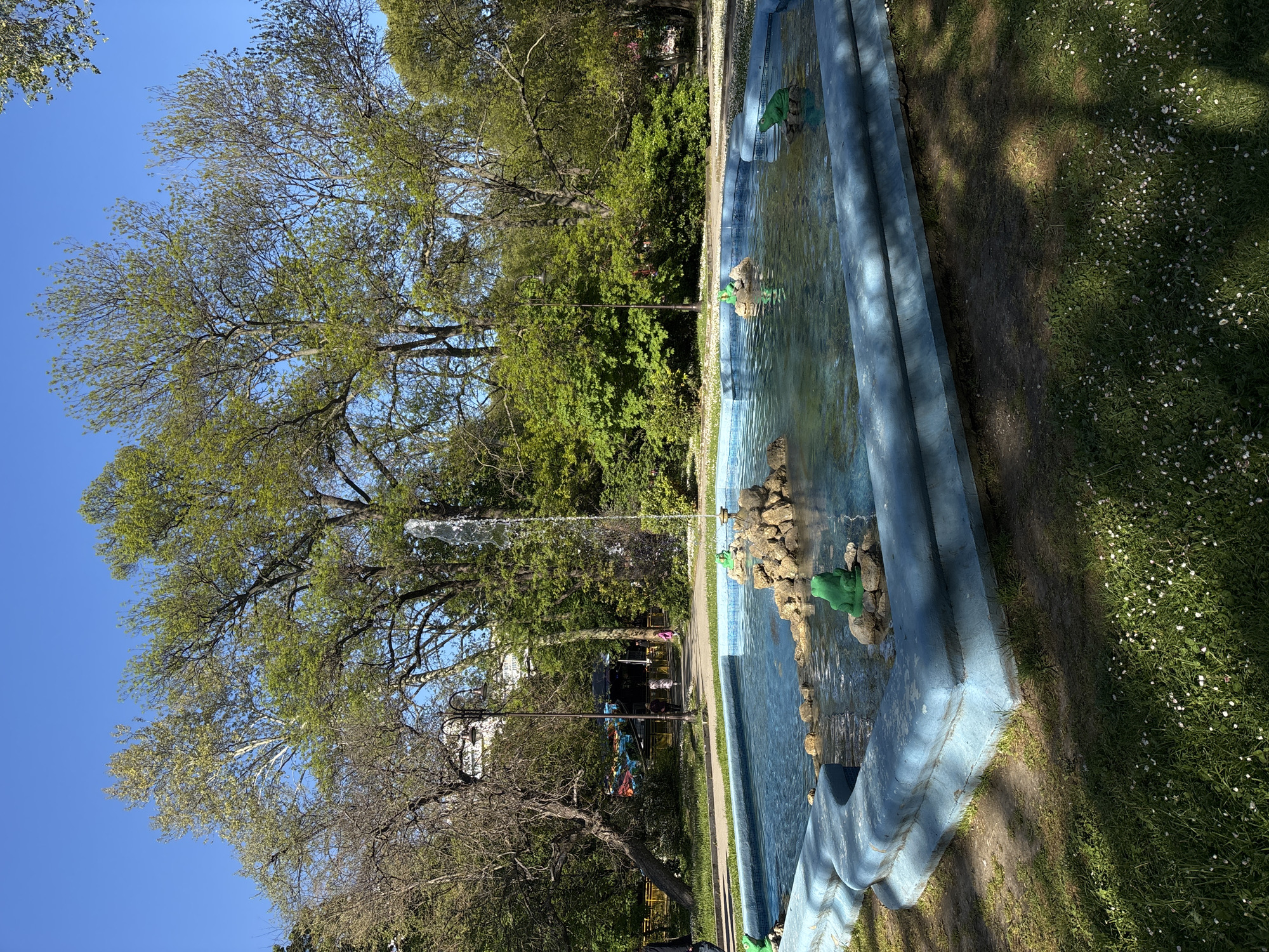 A long, light blue, rectangular fountain dominates the right side of the image, oriented toward the viewer. The fountain is filled with water and features multiple tiers and fountainheads, some adorned with green frog sculptures. Three water jets are visible spraying water upward.
To the left of the fountain, a park setting unfolds with lush green trees and grass. A walking path runs parallel to the fountain, and in the distance, structures, possibly part of a park facility or buildings in the area, are visible. A lamp post stands along the path. The sky is clear and bright blue, indicating a sunny day.
Based on the presence of the distinctive fountain sculptures and overall park setting, the image likely depicts a scene in the Sea Garden (Morska Gradina) park in Varna, Bulgaria.