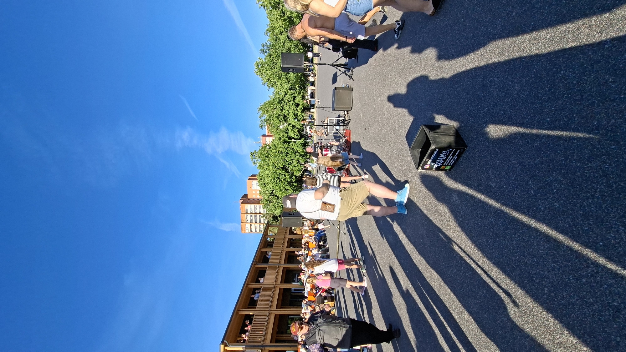 The image depicts an outdoor scene in Tallinn, Estonia, during a daytime event under a clear blue sky. The sun casts long shadows, indicating it is likely late afternoon or early evening. The setting appears to be an urban area, possibly a street or plaza, with a modern building featuring multiple levels of balconies on the left and mature trees lining the background.
The primary activity is a gathering with a band performing on a stage in the mid-ground. Several people are visible, some standing and observing the performance, while others are seated. A man in a white t-shirt and beige shorts is walking away from the camera, while a woman in a white top and denim shorts stands near the stage. In the foreground, a black box with text and logos is placed on the asphalt. The text on the box includes "BRADEE" in large letters, and other smaller text that appears to be social media handles and contact information.
The atmosphere seems relaxed and festive, with people enjoying the outdoor event. The presence of a band and the gathering of individuals suggest a concert, festival, or similar public event. The weather is sunny and pleasant, contributing to the outdoor ambiance. The architecture of the building and the general urban landscape suggest a European setting.
