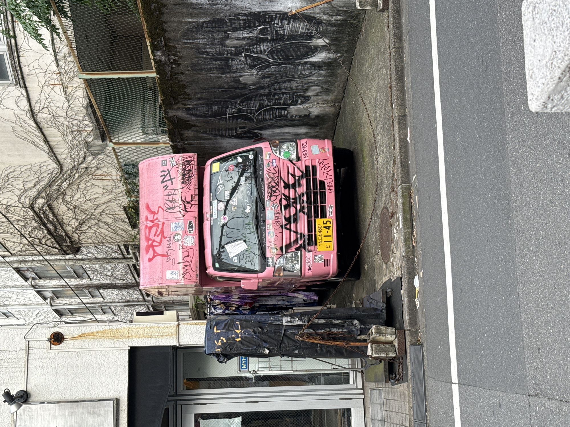 In Shinjuku, Japan, a pink delivery truck is parked on a concrete surface adjacent to a building. The truck is heavily adorned with graffiti, both on its exterior panels and on the roof cargo box. The surrounding environment includes a building wall covered in graffiti, a chain-link fence with moss, and a street in the foreground with a white lane marker. To the left of the truck, a black, tarpaulin-covered object stands on a wheeled platform, also showing signs of graffiti and wear. The building behind the truck has a white facade with a prominent vine growth.