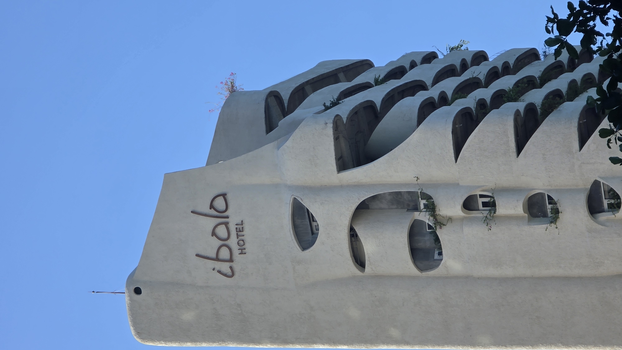 Viewed from below and angled, the picture depicts the exterior of the "¡bala HOTEL" building in Bac Nha Trang Ward, Vietnam. The structure is made of light-colored concrete, featuring an irregular design with rows of repeating arch-shaped openings at the top, some containing plants. A series of oval-shaped openings can be seen on the lower portion of the building, some of which also have plants growing out of them. The hotel's name is vertically inscribed on the side of the building in a dark, sans-serif font. The background consists of a clear, light blue sky, with some foliage visible on the right side of the frame.