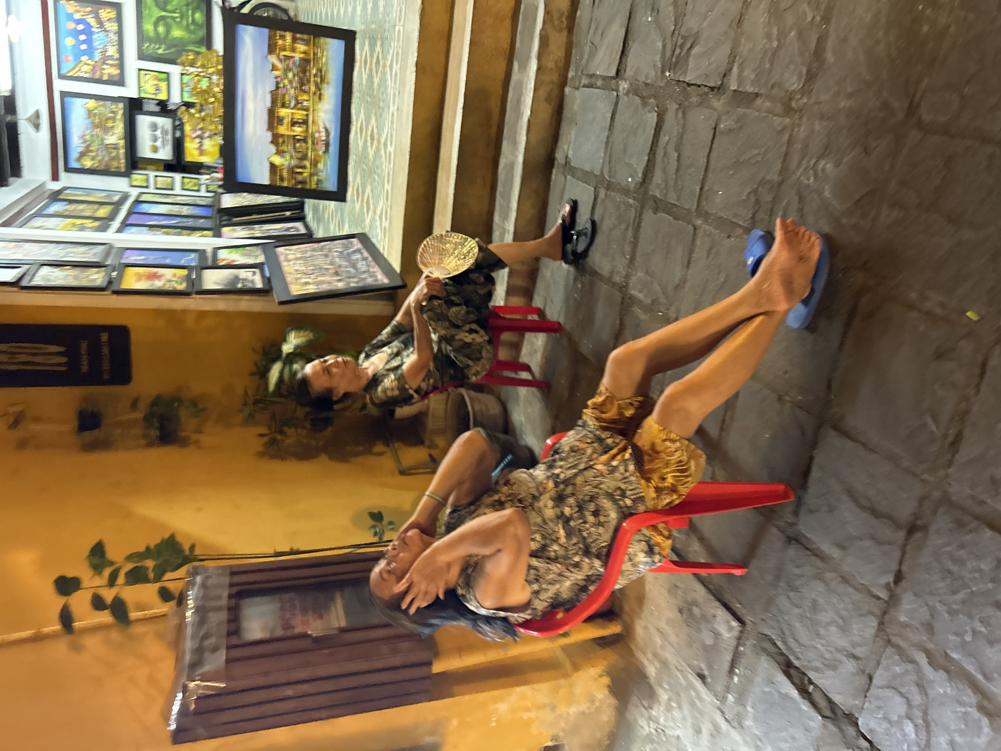 In Hội An Ward, Vietnam, two women sit on red chairs outside an art shop. The woman on the left, wearing a patterned top and yellow shorts, holds her hands to her head. The woman on the right, in a patterned dress, holds a fan. The shop displays numerous paintings on its walls and leans several more against its exterior. The ground is paved with irregular stone tiles.