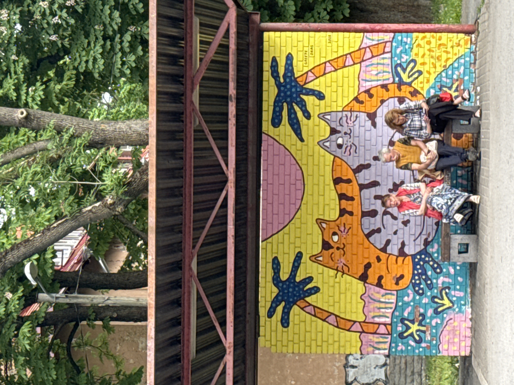 Three individuals, two women and one man, are seated on a bench beneath a shelter. The bench is positioned in front of a vibrant mural painted on a brick wall. The mural depicts two large, stylized cats in shades of orange and grey, surrounded by palm trees and a colorful, patterned background. The woman on the left wears a red vest over a patterned dress and a pink scarf. The man in the middle is dressed in a yellow vest over a light-colored shirt and dark pants. The woman on the right wears a plaid shirt and black pants, and is looking at her phone. The shelter above them has a corrugated metal roof supported by a red metal frame. Trees with green foliage are visible behind the shelter. The ground in front of the bench appears to be paved. The mural has signatures and a date that reads "2026" which is likely the year the mural was painted, though it is a future date. The context provided indicates this scene is in Tallinn, Estonia.