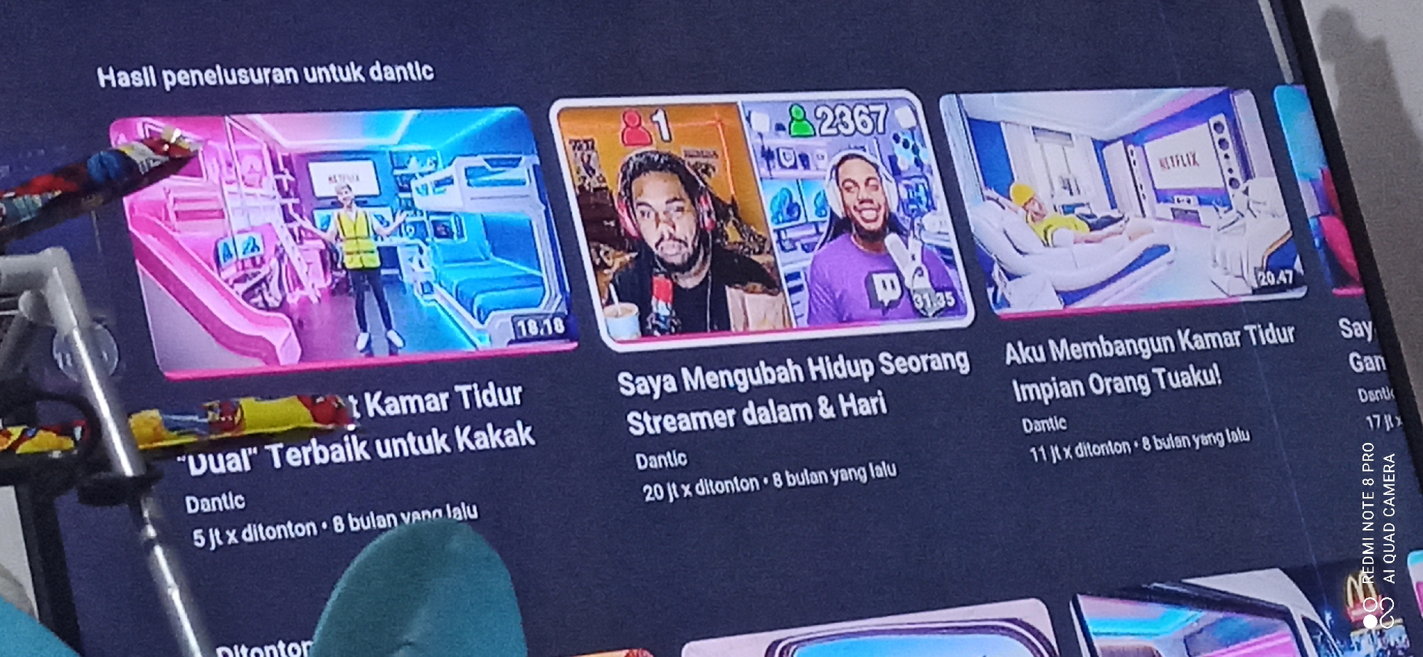 The image displays a smartphone screen showing a YouTube interface with several video thumbnails and text. The videos appear to be from a creator named "Dantic". One video title is ""Uual" Terbaik untuk Kakak" with 5 million views. Another video is titled "Saya Mengubah Hidup Seorang Streamer dalam & Hari" with 20 million views. A third video is titled "Aku Membangun Kamar Tidur Impian Orang Tuaku!" with 11 million views. The videos are presented in a vertical layout typical of mobile YouTube. The phone is placed on a surface, and a portion of a person's hand holding the phone is visible on the left edge of the image. The context of Sedati Agung, Indonesia is not directly discernible from the visual information provided in the image itself.