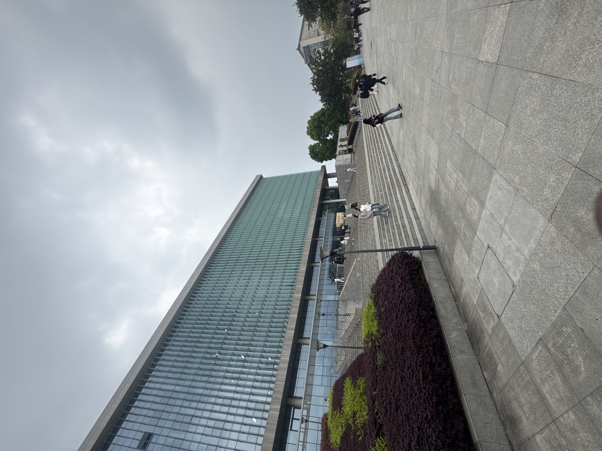 This image depicts a modern building with a glass facade located in Jiangjin, China. The building is a tall rectangular structure, possibly an office building or university building, with a linear pattern of windows. In front of the building, there is a paved area with large gray tiles and steps leading up to the entrance. People are walking on these tiles. In the foreground, there is a long rectangular planter filled with bushes with maroon-colored leaves and some green foliage. The sky is overcast and gray.