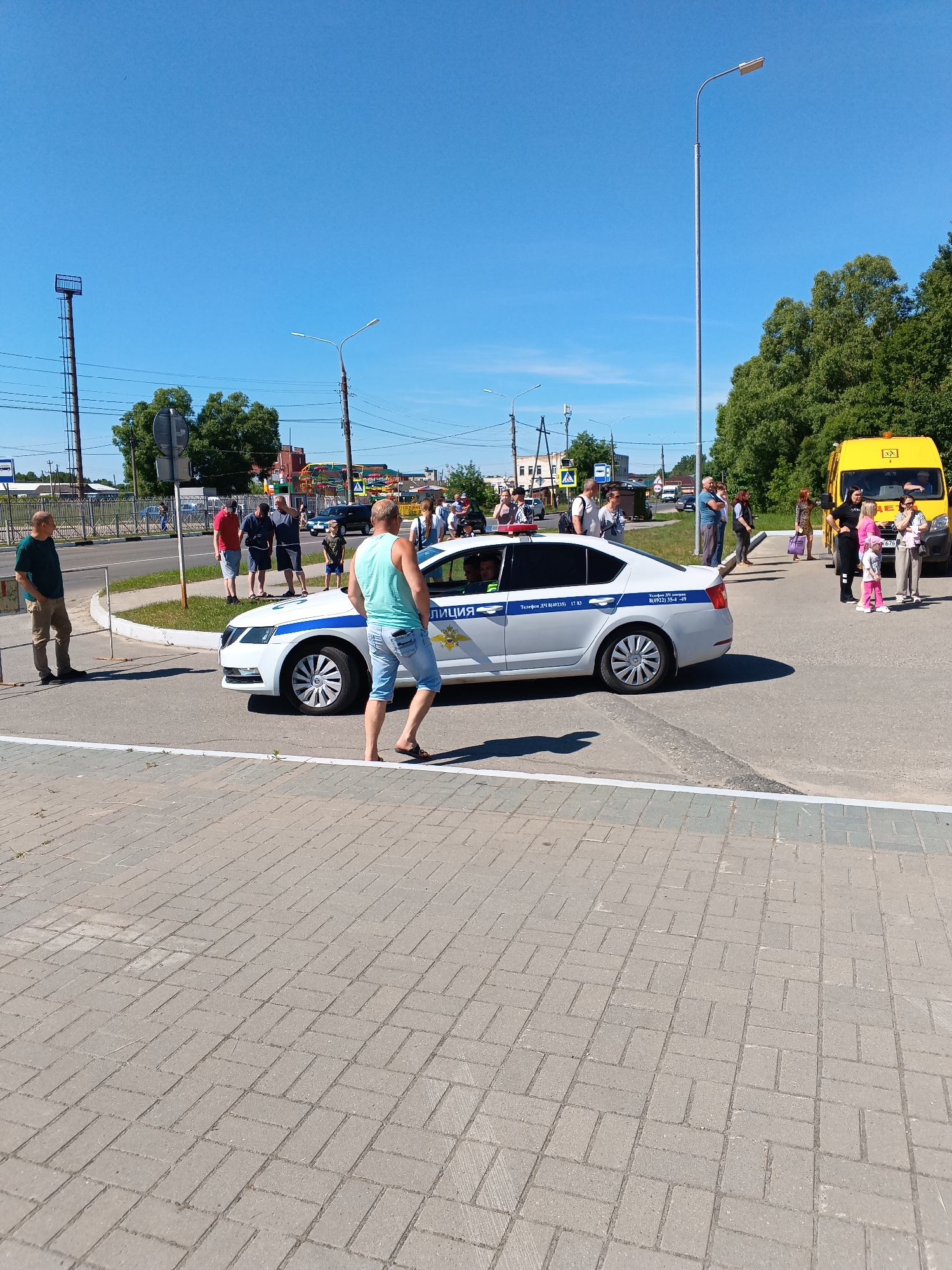 This image depicts an outdoor scene in Sudogda, Russia, under clear blue skies and bright sunlight, suggesting daytime. The primary focus is a white police sedan (likely a Skoda Octavia) with a blue stripe, displaying "ПОЛИЦИЯ" (Police) lettering and the emblem of the Russian Ministry of Internal Affairs (MVD) on its side. Red and blue emergency lights are mounted on the vehicle's roof. A person is visible in the driver's seat.
In the immediate foreground, a light grey sidewalk or pedestrian area with interlocking pavers laid in a herringbone pattern is visible. A man in a light blue tank top and denim shorts, holding a pair of flip-flops in his left hand, is walking away from the driver's side of the police car, his back partially to the camera. To his left, another man in a dark green t-shirt and tan pants stands with his hands in his pockets, observing the scene.
The police car is positioned on an asphalt road, angled towards the right side of the frame. A group of individuals, including men, women, and at least one child, are gathered around the car and further into the background. Many appear to be observing the vehicle or the general area. To the right of the police car, near a curb, a yellow van, resembling a common Russian "Gazelle" model, is parked. A small group of people, including a woman in black clothing with a young girl in pink, are standing near this yellow van.
In the mid-ground, a dark-colored sedan is visible driving on the road. Various street furniture is present, including tall metal street light poles with modern light fixtures, and multiple utility poles supporting overhead power lines. To the far left, a tall, dark metal tower with a small square platform at its top stands against the sky. Partially obscured by trees, two street signs are visible: a circular 'no entry' sign and a rectangular 'pedestrian crossing' sign. Lush green deciduous trees line the background, with distant buildings partially visible. The overall atmosphere appears calm, with people casually observing the scene.