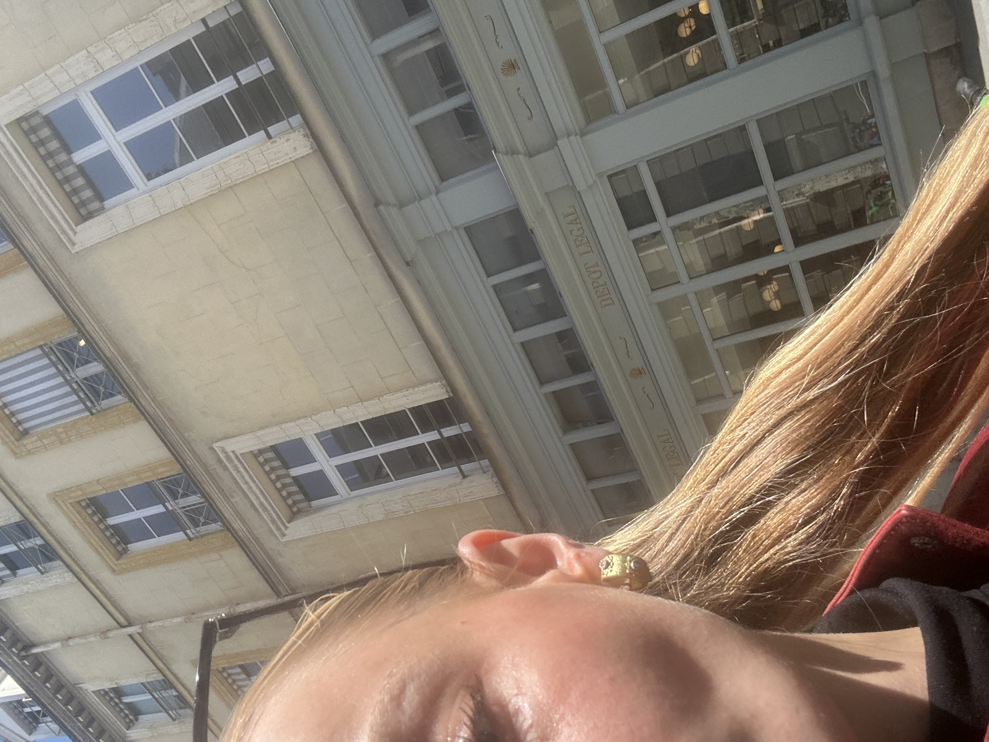 A distorted, low-angle, close-up photograph shows a person's face against a background of multi-story architecture. The person has light-colored hair and is wearing a gold earring. The building features numerous windows and architectural details, including the words "DEPOT LEGAL" inscribed on its facade. The location appears to be urban, with a building constructed with light-colored stone or concrete. The light in the image suggests it is daytime.