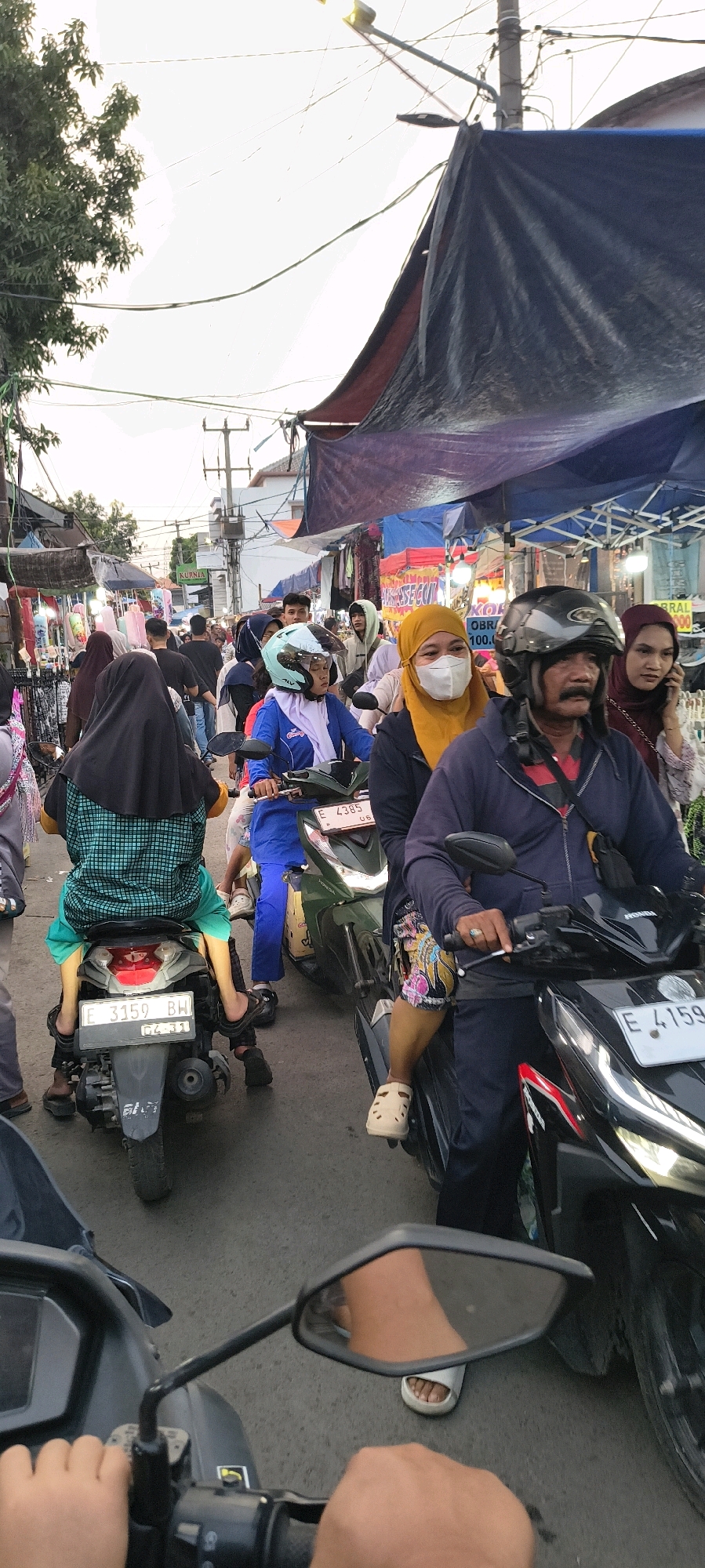 This is a vertical, eye-level, full shot of a busy street scene in Trusmi Wetan, Indonesia. The image captures a moment of traffic congestion, with numerous motorcycles and pedestrians navigating a narrow road lined with market stalls. The atmosphere is lively and crowded, indicative of a local marketplace or a festival day.
In the foreground, the perspective is from the rider of a motorcycle, with their hands visible on the handlebars and a portion of the motorcycle's console and mirror reflected in the frame. This viewpoint gives a sense of immediate immersion into the scene.
Dominating the mid-ground are several motorcycles carrying passengers, creating a sense of movement and density. One motorcycle in the center is ridden by a person wearing a blue jacket and a light blue helmet, with a person in a black and green checkered tunic riding behind. To their right, another motorcycle carries a person in a dark blue jacket and a helmet, with a passenger in a maroon headscarf. A woman in a bright yellow headscarf and a white mask is also visible on a motorcycle, her face turned slightly towards the viewer.
The background reveals a bustling market. Stalls with various goods are set up along the roadside, covered by tarpaulins. Colorful fabrics and items are displayed for sale. The presence of streetlights and electrical wires overhead suggests an urban or semi-urban setting. The overall lighting appears to be natural, possibly late afternoon or early evening, with a soft, diffused quality. The sky is mostly overcast or bright, with some trees visible on the left edge of the frame. The road surface is asphalt, showing signs of wear. The people in the scene are predominantly dressed in casual attire, with many women wearing headscarves, which is common in Indonesia. The overall impression is one of a vibrant and active street life in a commercial area.