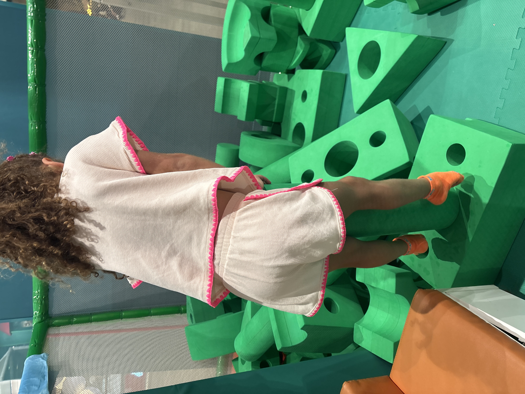 A young girl with curly brown hair, wearing a white waffle-knit outfit with pink trim and orange socks, stands on a stack of bright green foam blocks in a soft play area. The background features a green safety net enclosure and a light blue wall. The girl is facing away from the camera and appears to be climbing or exploring the soft play structures. The scene is likely located within an indoor play center in Dubai, United Arab Emirates, given the context provided.