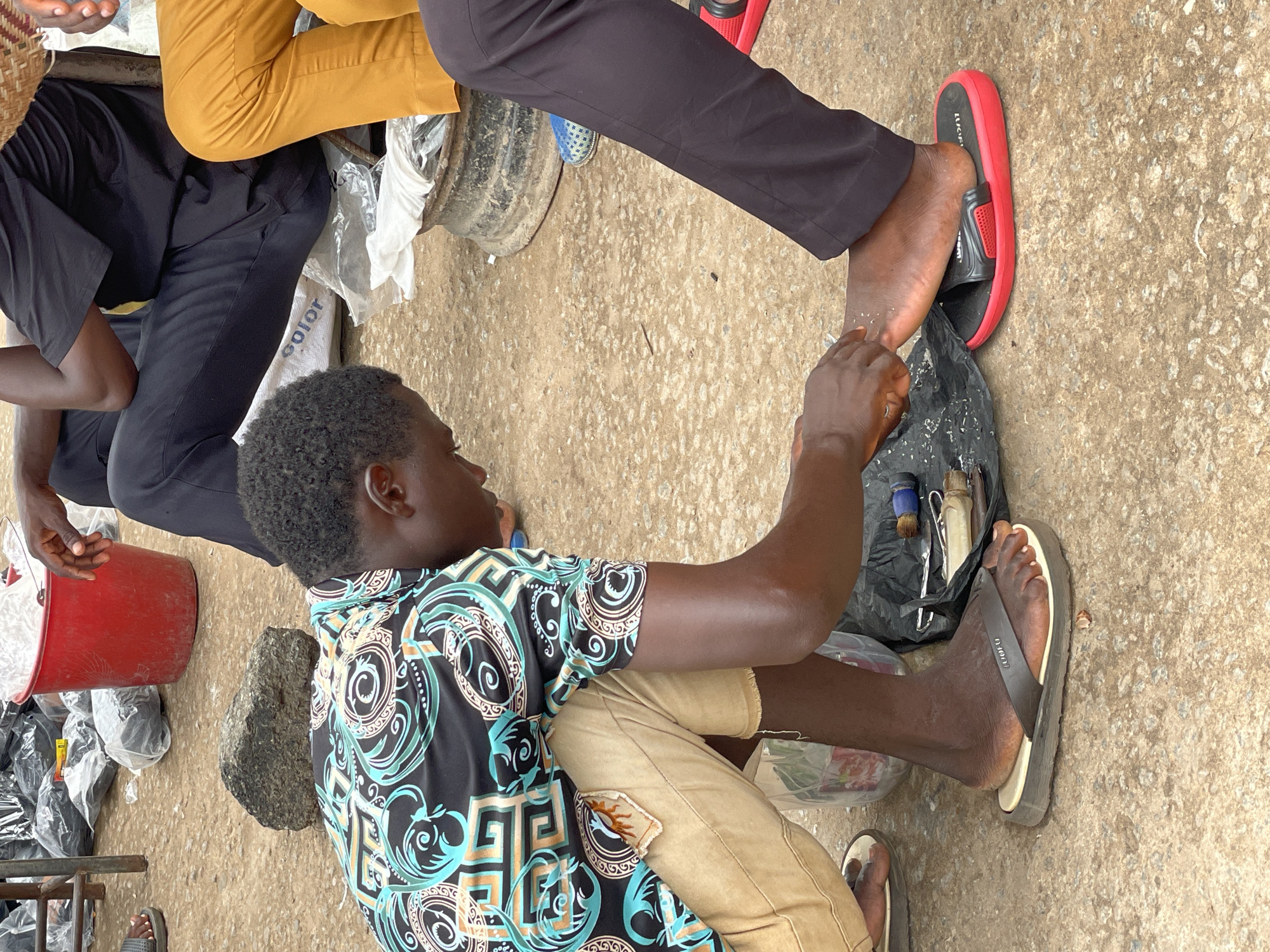 In the city of Onitsha, Nigeria, an outdoor scene unfolds on a concrete ground surface. A man is seated, wearing a black and teal patterned short-sleeved shirt and khaki pants. He is working on another individual's foot, which is adorned with a red and black sandal. A black plastic bag containing tools rests beside him. Next to the seated man is a red bucket and bags of what appear to be sandals. Adjacent to the foot care activity, two other individuals are present: one wears yellow pants, and the other black pants.