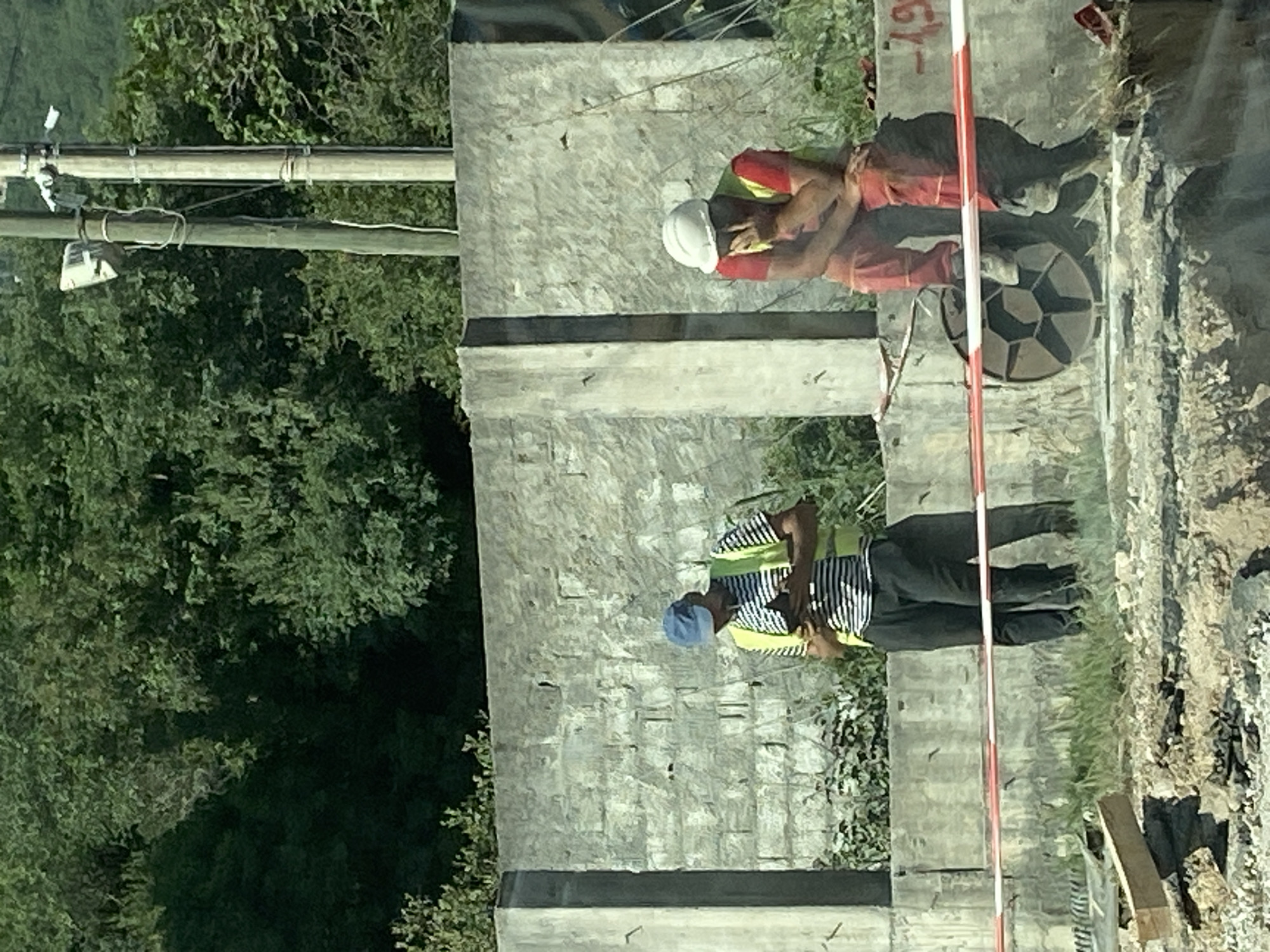 The image depicts a construction site in Lastva Grbaljska, Montenegro. Two construction workers are visible. One worker wears a white hard hat, red shirt, and red pants. The other worker wears a blue cap, a striped shirt, a neon yellow safety vest, and dark pants. They are standing on or near a concrete structure. Nearby is a utility pole with wires and a small, attached device. A red and white striped pole is positioned as a barrier. The environment includes dense green trees. There is a black manhole cover.