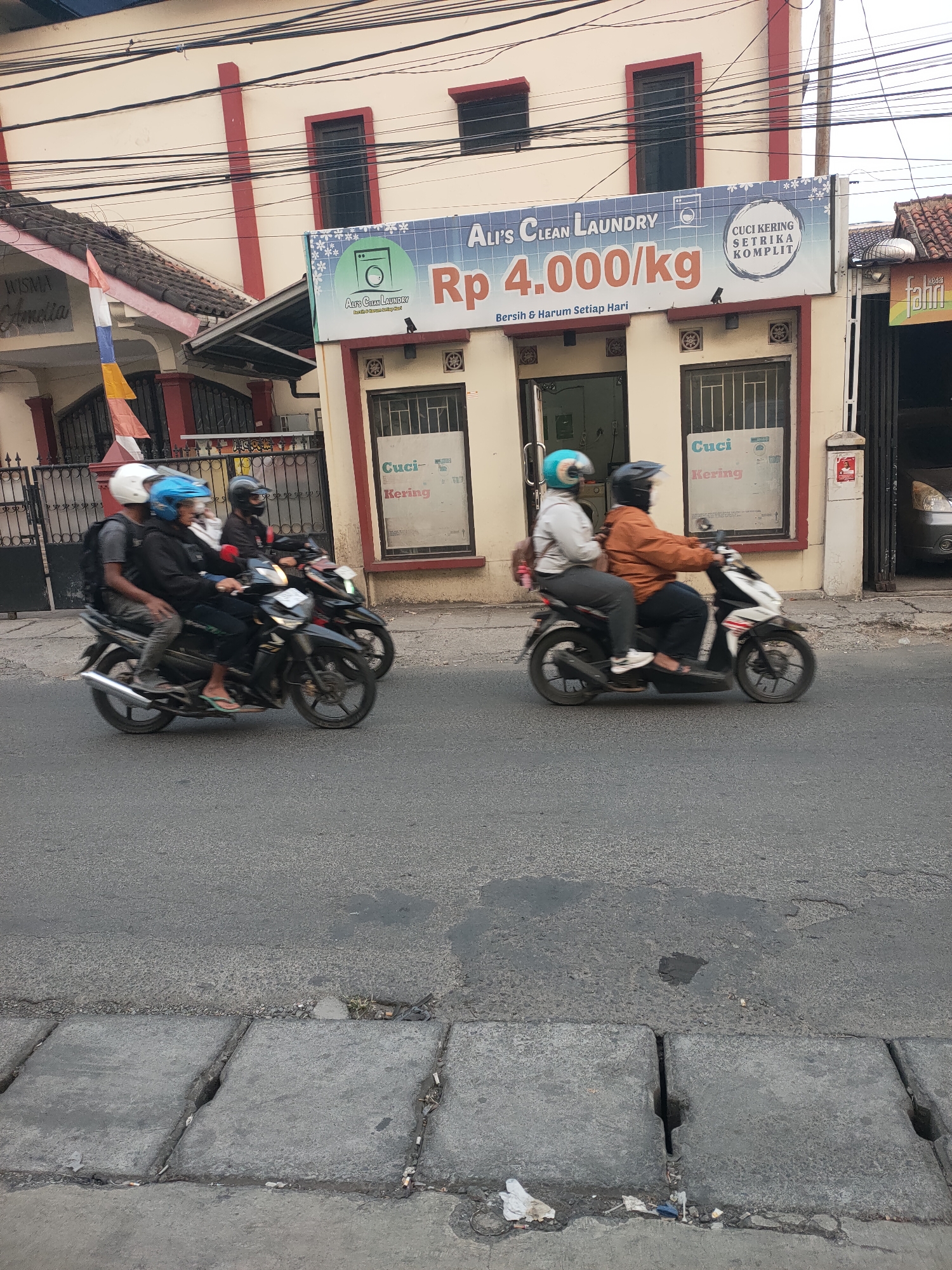 This image captures a street scene in Cimahi, Indonesia, with a laundry service visible in the background. In the foreground, two motorcycles are in motion. The first motorcycle, on the left, is carrying three individuals. The rider is wearing a black helmet. The passenger behind the rider has a blue helmet, and the person seated behind them is wearing a white helmet. The second motorcycle, on the right, has two occupants. The rider is wearing an orange jacket and a dark helmet, while the passenger is wearing a light-colored jacket and a blue helmet.
The background features a building with a sign for "Ali's Clean Laundry." The sign advertises laundry and dry cleaning services with a price of Rp 4,000/kg. Below the main sign, smaller signs in Indonesian indicate "Cuci Kering Setrika Komplit" (Complete Dry Cleaning and Ironing). The building itself appears to be a commercial establishment with windows and doorways. The street is paved, and a concrete drainage system runs along the bottom of the frame. Overhead, a tangle of electrical wires is visible. The overall environment suggests a typical urban street in Indonesia.