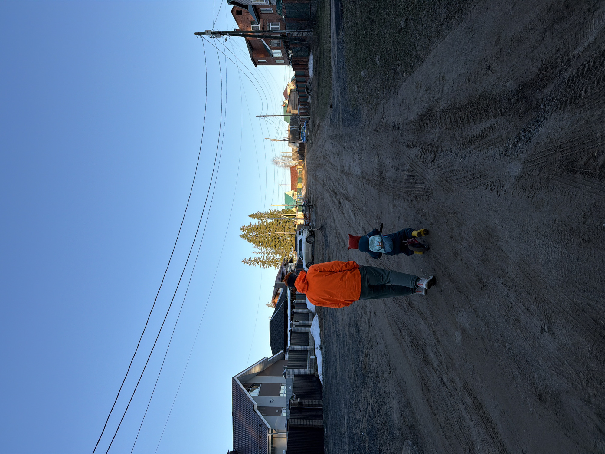 A slightly rotated long shot shows a man and a small child walking down a dirt road in Chaikovsky, Russia. The man, facing away from the viewer, is wearing an orange jacket and green pants. The child is wearing dark clothing and has a backpack. They are walking on the right side of the road, which is heavily textured with tire tracks. Houses line both sides of the road, and a utility pole with wires is visible above. In the distance, trees and a white car can be seen. The sky is clear and blue.