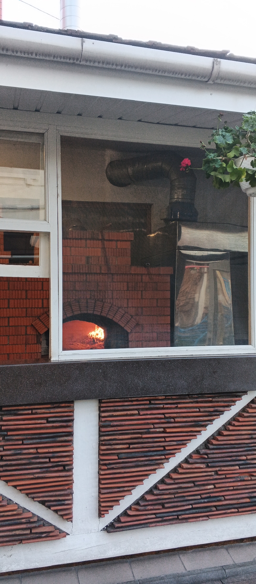 The image shows a brick pizza oven with flames visible inside, viewed through a window. The oven is built into a wall of bricks and has an arched opening. Above the oven, a metal pipe extends upwards and out of view. A hanging plant with a pink flower is visible in the upper right corner. The exterior of the building features a patterned brick facade below the window. The location is identified as Berehove, Ukraine.