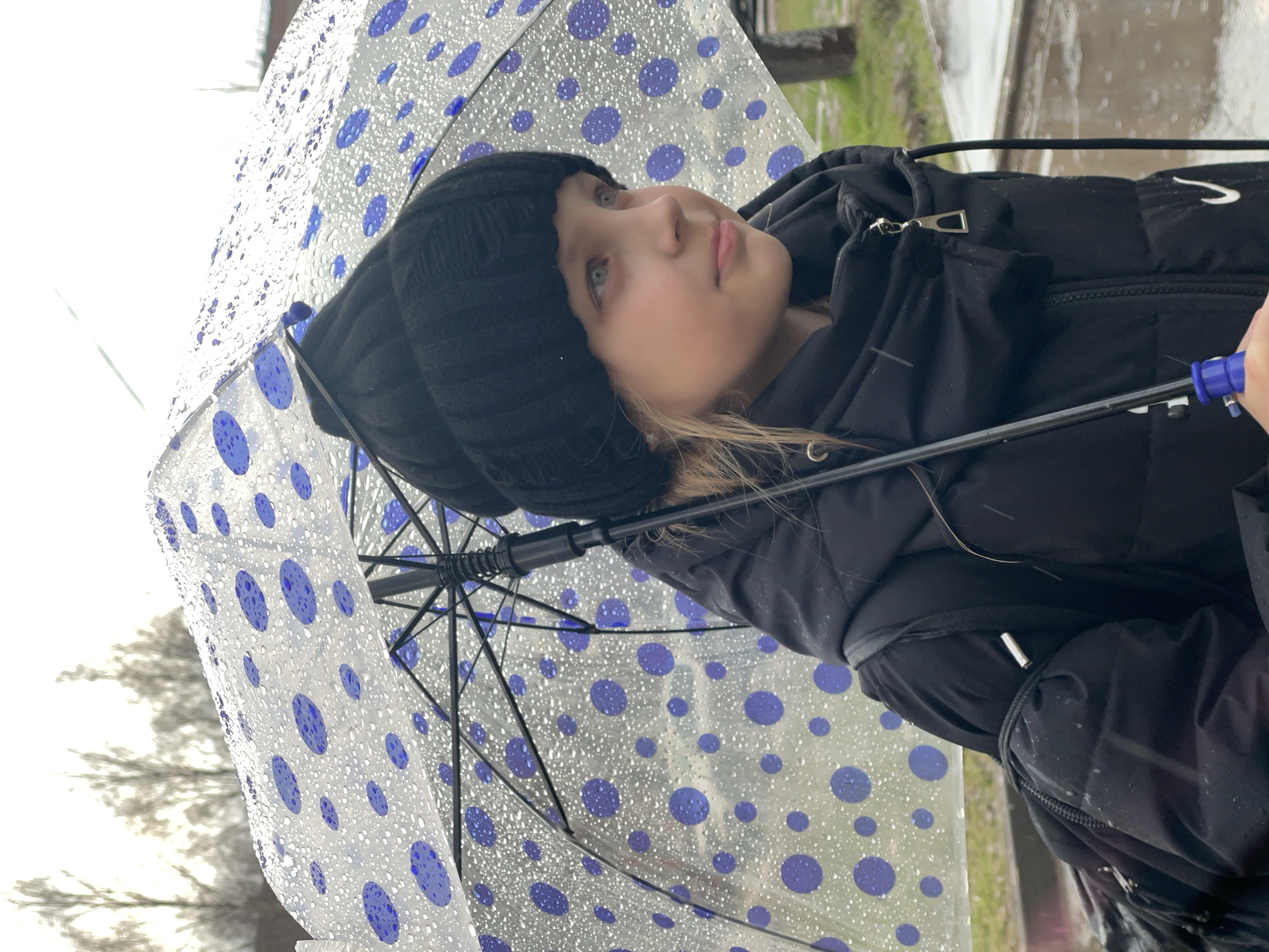 A girl stands outside holding a transparent umbrella with blue polka dots. She wears a black knit hat and a black puffer jacket. The background is blurred, suggesting an outdoor location. Specific details indicating Izhevsk, Russia, are not discernible from the visual information.