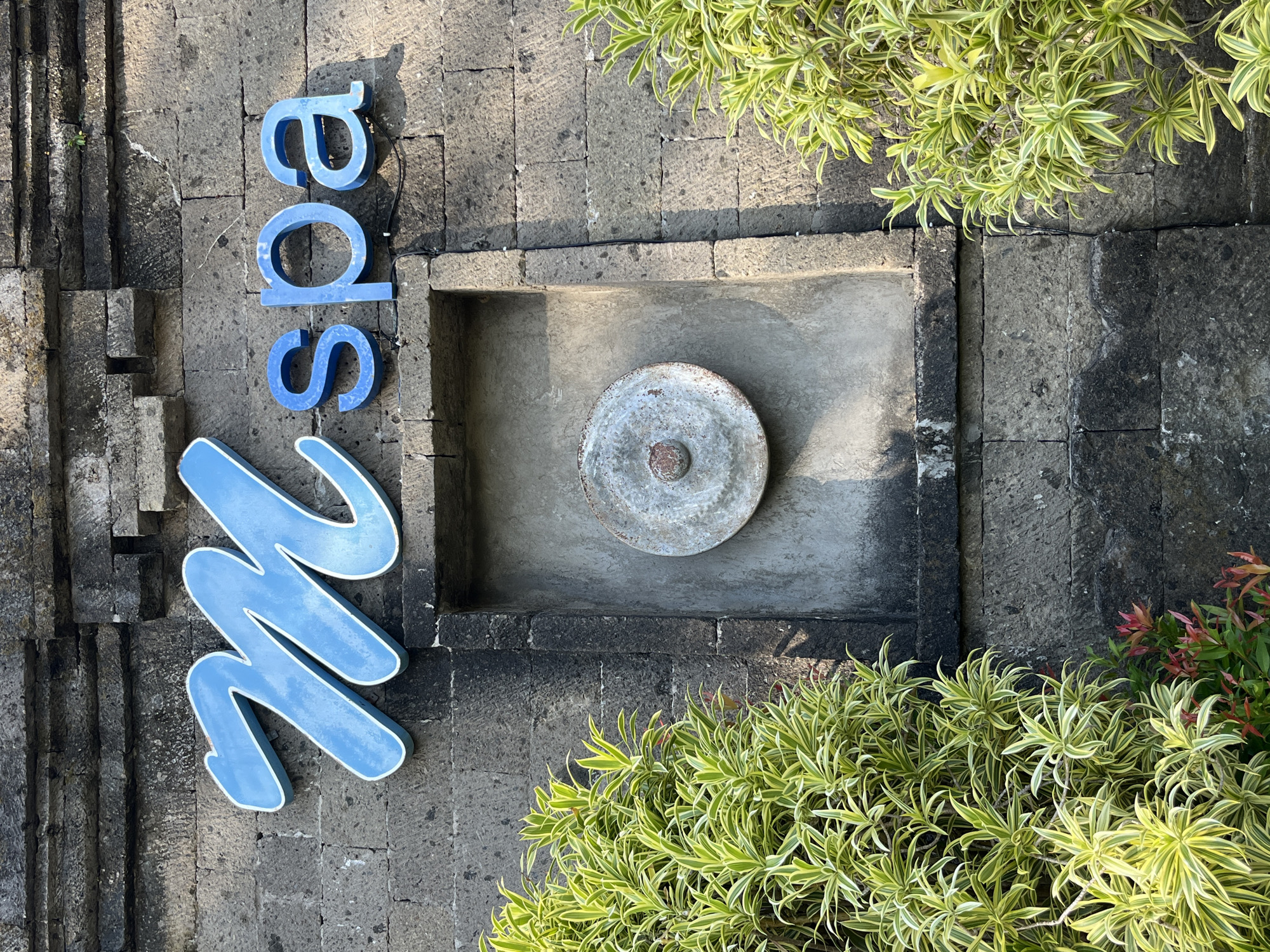 In Kuta Selatan, Indonesia, a stone wall displays the blue 3D letters "M spa." The letters are outlined in white and appear to be mounted on the wall. To the left and right of the signage, lush green plants with variegated leaves frame the scene. In the center of the wall, behind the "M spa" lettering, a recessed rectangular area contains a round, metallic object, possibly a gong or a decorative piece, with a central protrusion. The stonework of the wall is rough-hewn and appears aged. Sunlight casts shadows on the wall, indicating an outdoor setting.