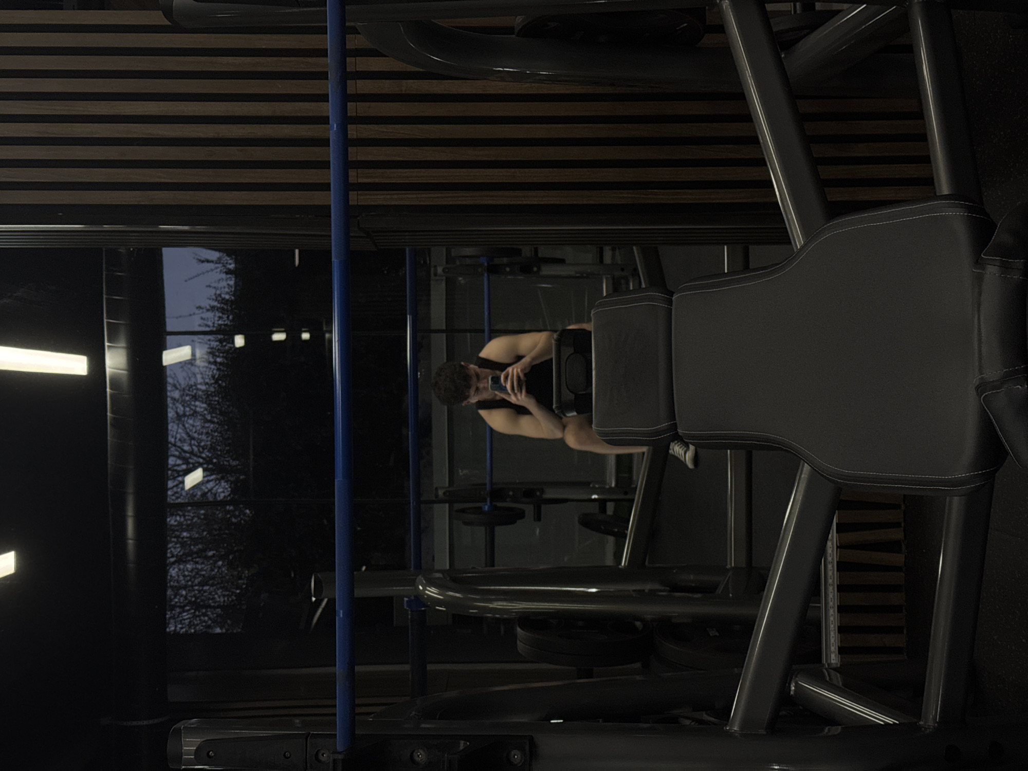 The image shows the interior of a gym, likely in Sofia, Bulgaria, as requested. The scene is dimly lit, with light sources visible through a large window at the left. The window appears to show bare tree branches silhouetted against a dusk or night sky.
Foreground elements feature a large exercise machine. The machine's seat is prominent, upholstered in dark grey and stitched with white thread. The frame of the machine is dark grey metal.
In the middle ground, a man is seated on another exercise machine, facing away from the primary machine in the foreground. He is holding a cell phone, seemingly taking a photo or using the device.
The gym's interior details include a wall with horizontal wooden slats above the windows and other gym equipment in the background, indicating a professional fitness environment. Weight plates are attached to the machines.
