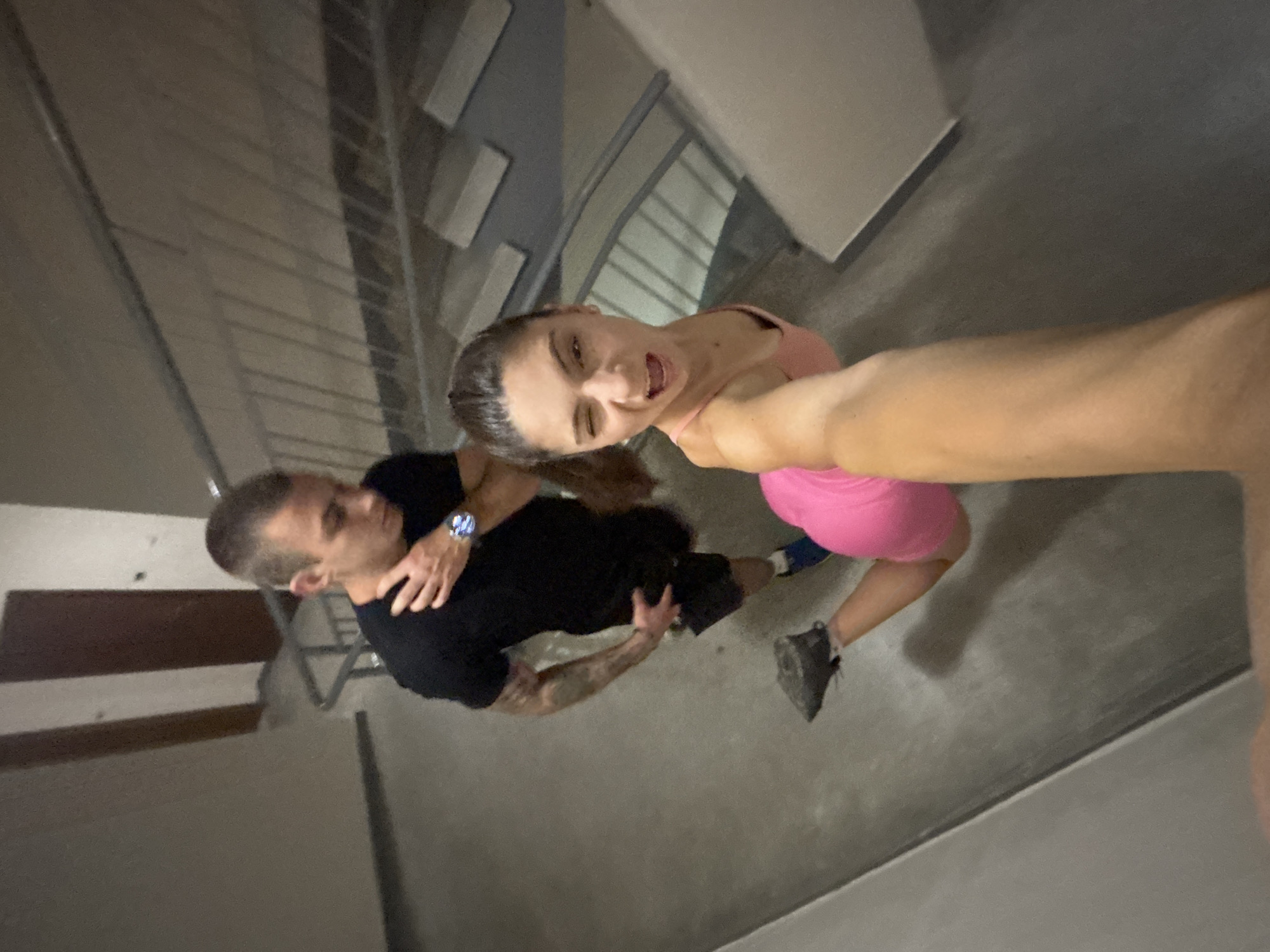In the city of Varna, Bulgaria, a young couple is captured in a selfie on a concrete staircase. The woman, positioned in the foreground and slightly to the right, is leaning into the shot with her right arm extended, holding the camera. She is wearing a bright pink tank top and pink shorts, with her left leg bent at the knee and her left foot resting on the stair below her. She is winking and has an open-mouthed expression, suggesting playfulness. The man stands behind her and to her left, looking down and to his left. He is wearing a black t-shirt and black shorts, with visible tattoos on his left arm and shoulder. He appears to be touching his chest or collarbone with his right hand, and his left hand is near his hip. The staircase has gray concrete steps and a metal railing. The overall environment suggests an indoor public space, possibly a building's stairwell or an outdoor staircase in a communal area.