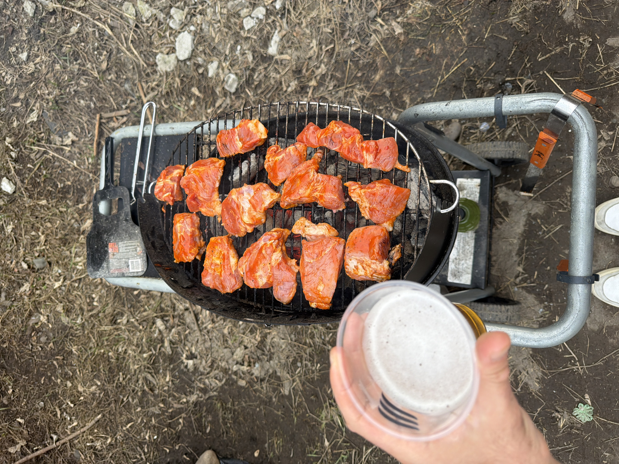 A top-down view captures a barbecue scene. Raw, marinated pieces of meat rest on a circular grill over hot coals. The grill is part of a portable, wheeled barbecue. The surrounding ground is covered in dry leaves and twigs, with scattered rocks. A person holds a clear plastic cup filled with a light-colored beverage, possibly beer or cider, near the grill. The scene is likely outdoors, given the natural environment. There is no information available to confirm that the image was taken in Krasnoyarsk, Russia.
