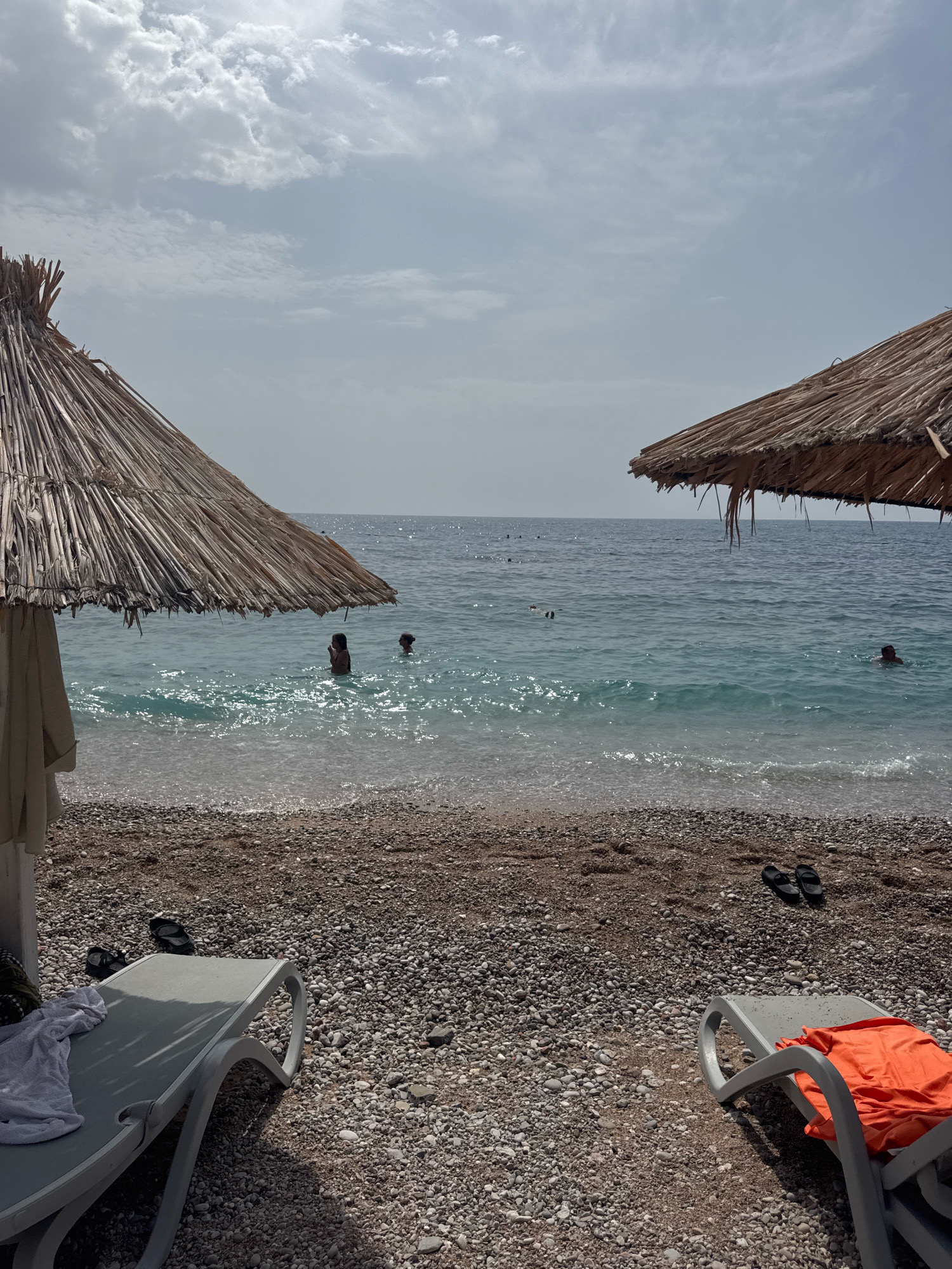 The image depicts a serene beach scene in Mirište, Montenegro. Two thatched umbrellas provide shade over a pebble beach. In the foreground, two empty lounge chairs are positioned on the pebbles, one with a white towel draped over it and the other with an orange towel. Several pairs of flip-flops are scattered on the beach. The turquoise waters of the Adriatic Sea gently lap at the shore. In the middle ground, several individuals are visible swimming and enjoying the water. The sky is partly cloudy, with a mix of sunlight and diffused light creating a soft ambiance. The overall impression is one of a tranquil day at the beach.
