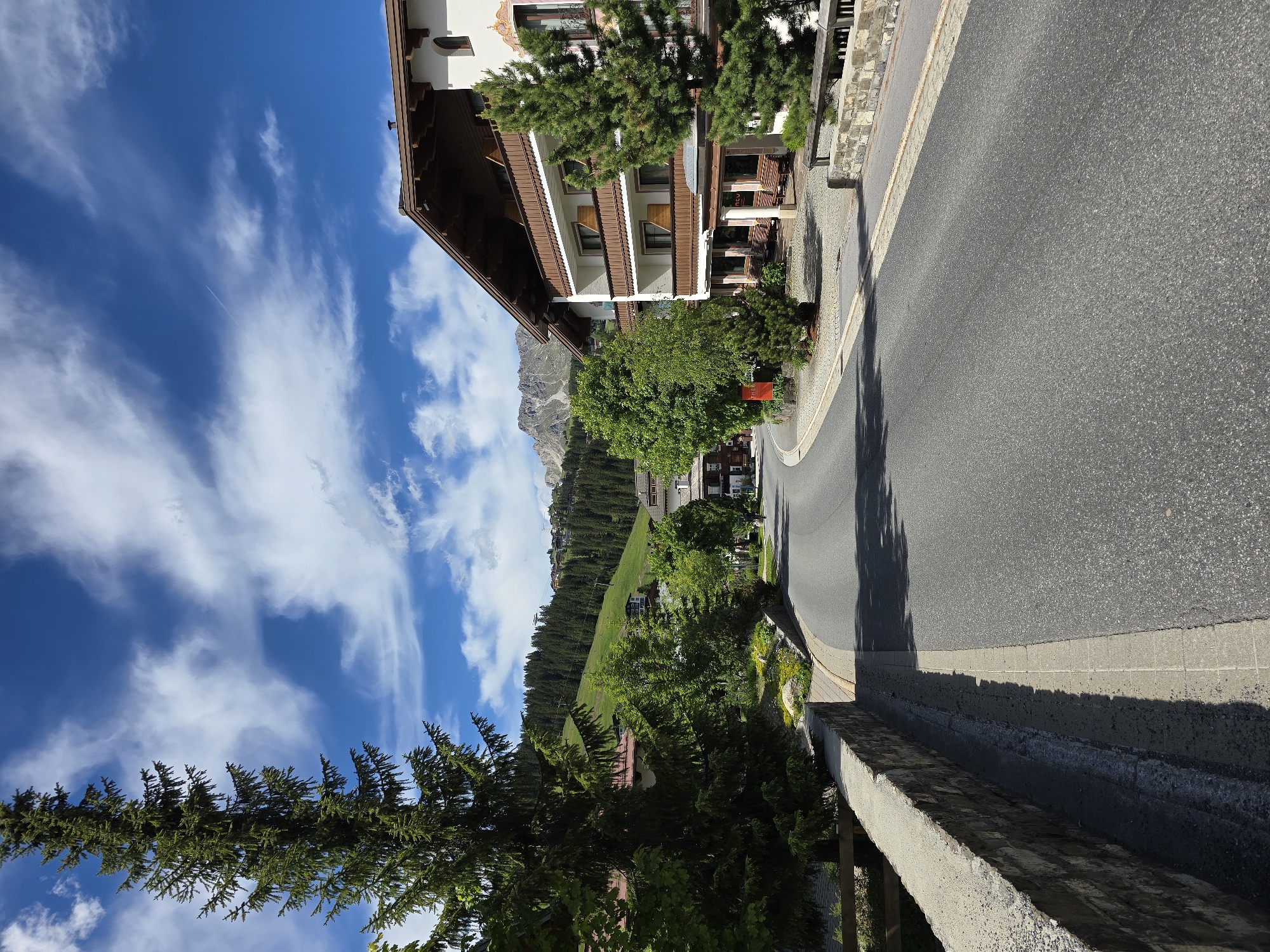 The image shows a paved road curving through a village with traditional Austrian architecture. On the right side of the road is a multi-story building with a brown wooden roof and balconies. Trees and greenery are abundant throughout the scene. In the background, a forested mountain slopes upwards, with some scattered houses and what appear to be ski lifts visible on its higher slopes. The sky is blue with white, wispy clouds. The location is identified as Lech, Austria. There are no people visible in the image. The overall impression is of a scenic mountain village in good weather.