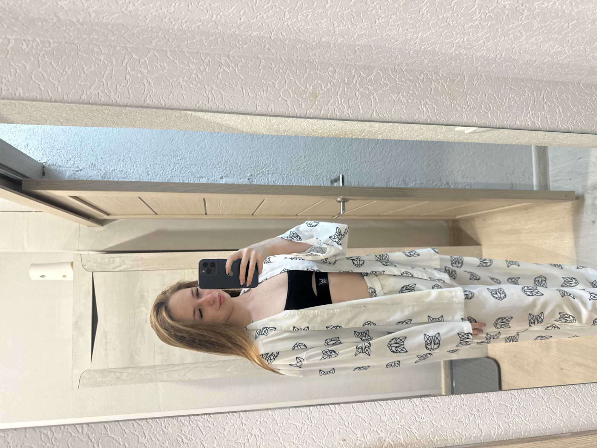 A young woman with long, light brown hair is standing in front of a mirror, taking a selfie with her phone. She is wearing a white pajama set with a black cat pattern and a black sports bra. The background shows a room with a wooden door and textured walls.