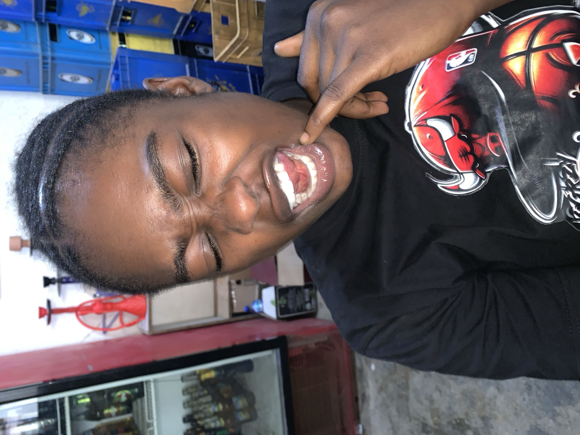 A person with dark skin, wearing a black shirt with a Chicago Bulls logo, is opening their mouth wide and pointing at their upper lip. They have their eyes closed and appear to be in some discomfort. Behind the person, there is a cooler with glass doors displaying drinks, blue crates stacked on top of one another, and various items on a shelf. This location is in the city of New Stadium, Nigeria.