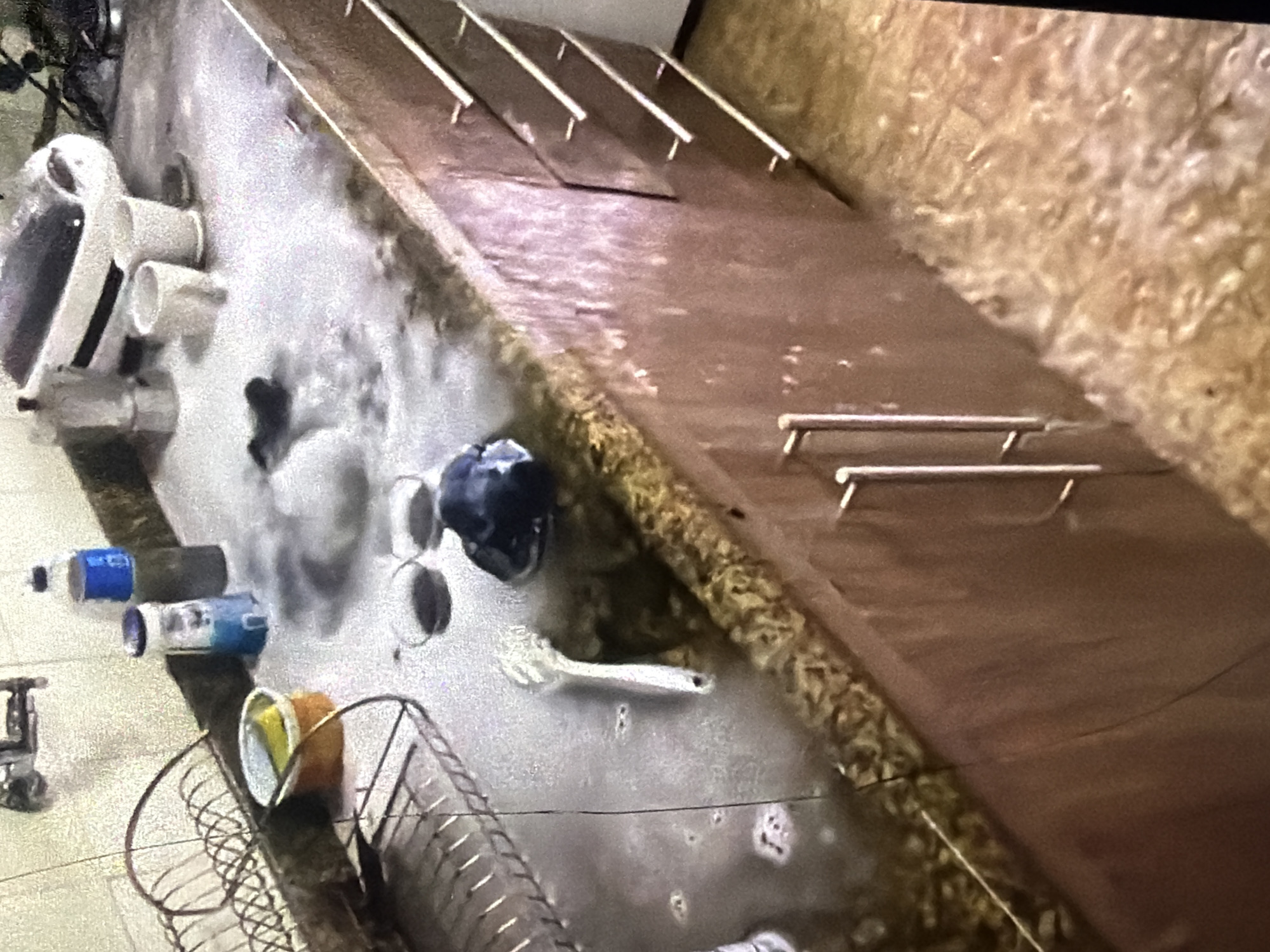 The image shows a kitchen counter overflowing with suds, indicating a potential plumbing issue or excessive use of cleaning products. Several objects are visible on the counter, including a coffee maker, cups, bottles, a sponge in a dish, and a spoon. The cabinets below the counter are a dark wood color with silver handles. The floor appears to be tiled. The scene suggests a domestic environment in Bukuru, Nigeria, where a clean-up or problem-solving situation is likely occurring.