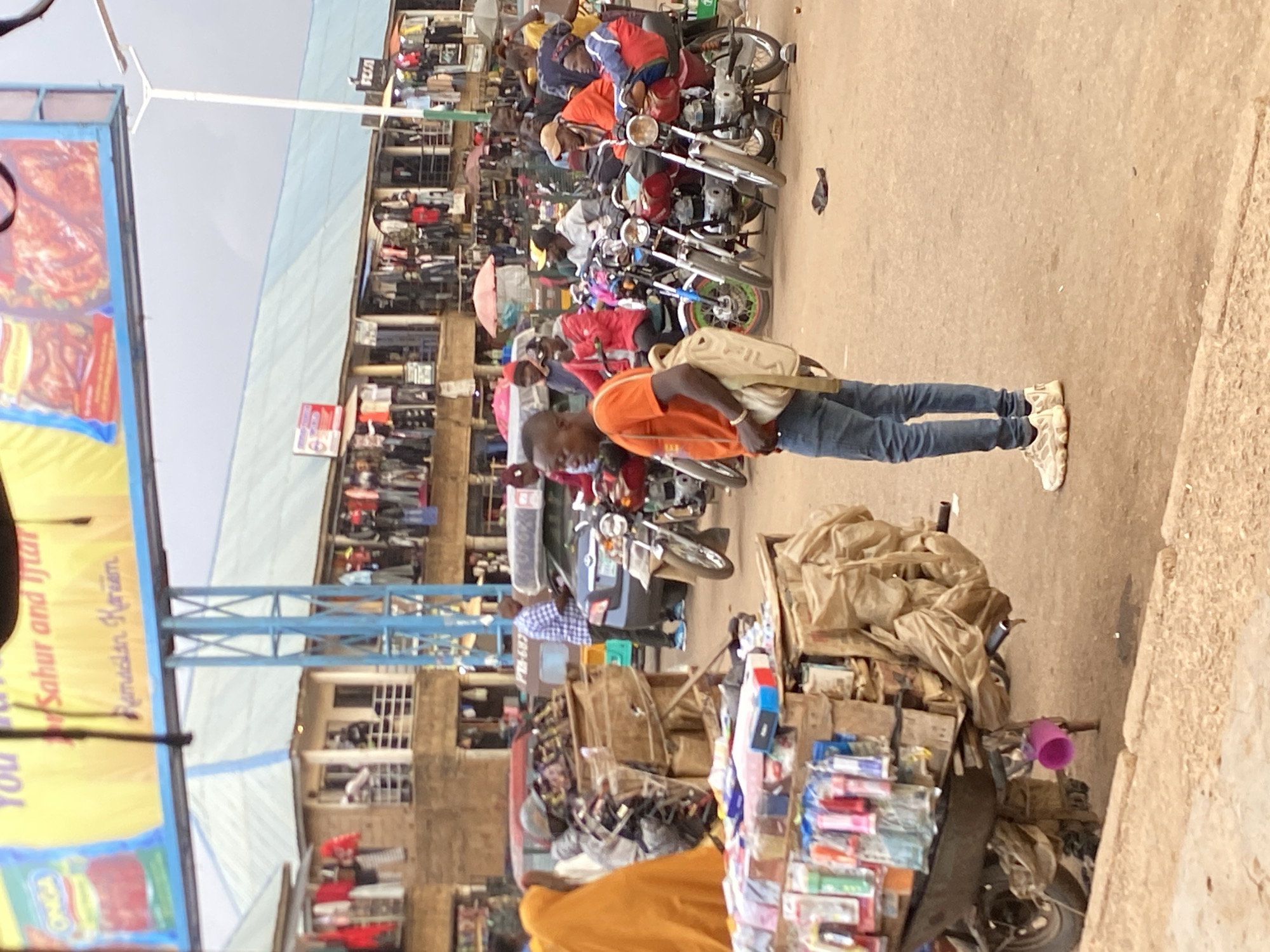 The image depicts a street scene in Bukuru, Nigeria. A man wearing an orange shirt and blue jeans stands on the right side of the frame, near a motorcycle piled with goods for sale. In the background, a busy street features a row of motorcycles parked along the side and a multi-story building with shops and stalls. A large sign is on the left side of the frame. The ground is paved.