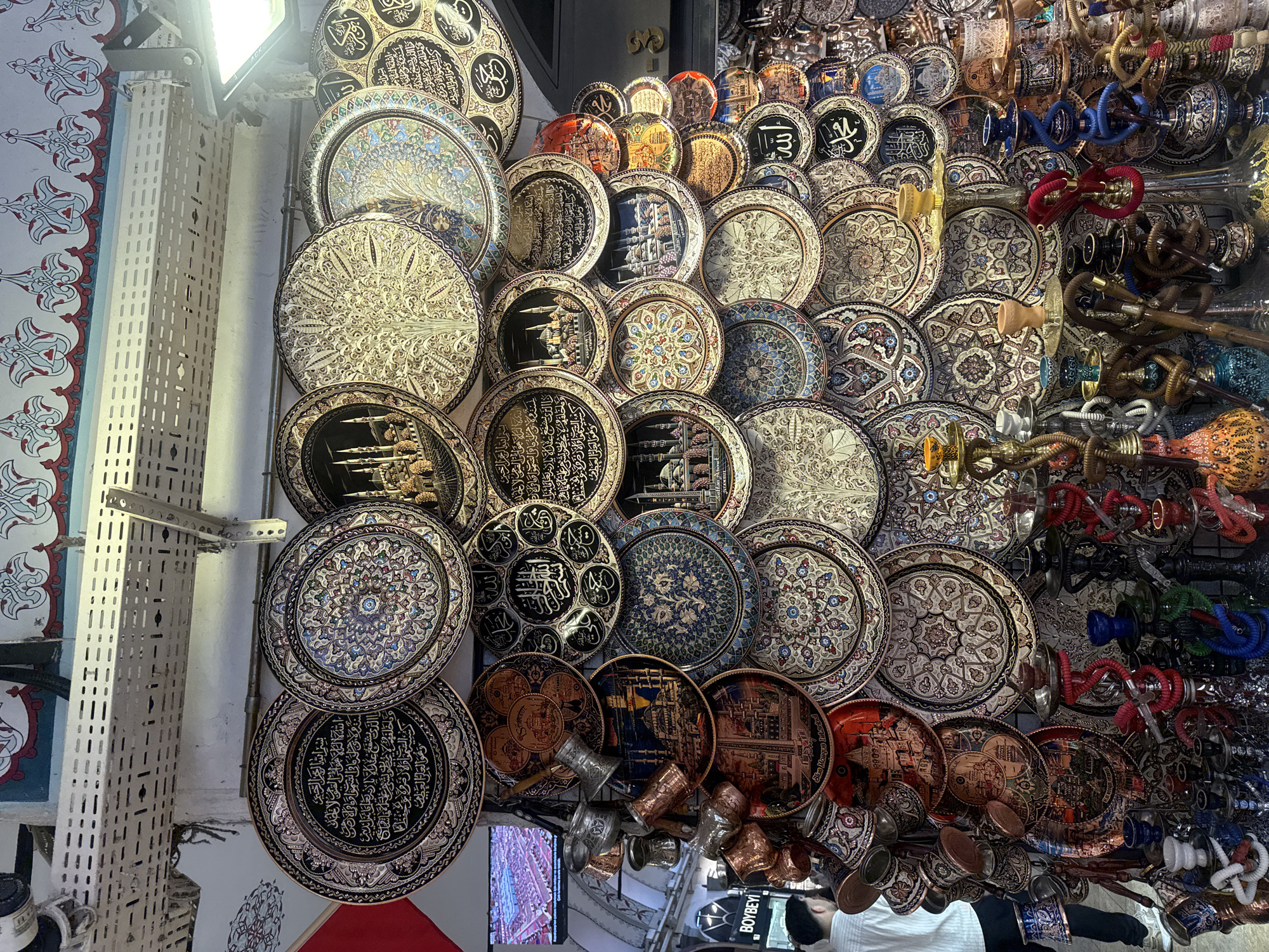 The image showcases a vendor stall in a market, likely located in Turkey based on the items displayed, filled with intricately designed metal plates, decorative cups, and hookahs. The plates are adorned with various patterns, including floral motifs, ships, and Islamic calligraphy. The hookahs are arrayed in a variety of colors and designs. Some cups are also displayed. A glimpse of a television screen and a man wearing a shirt can be seen in the bottom center. The location is potentially Istanbul based on the text "Blue Mosque Istanbul" seen on one plate.