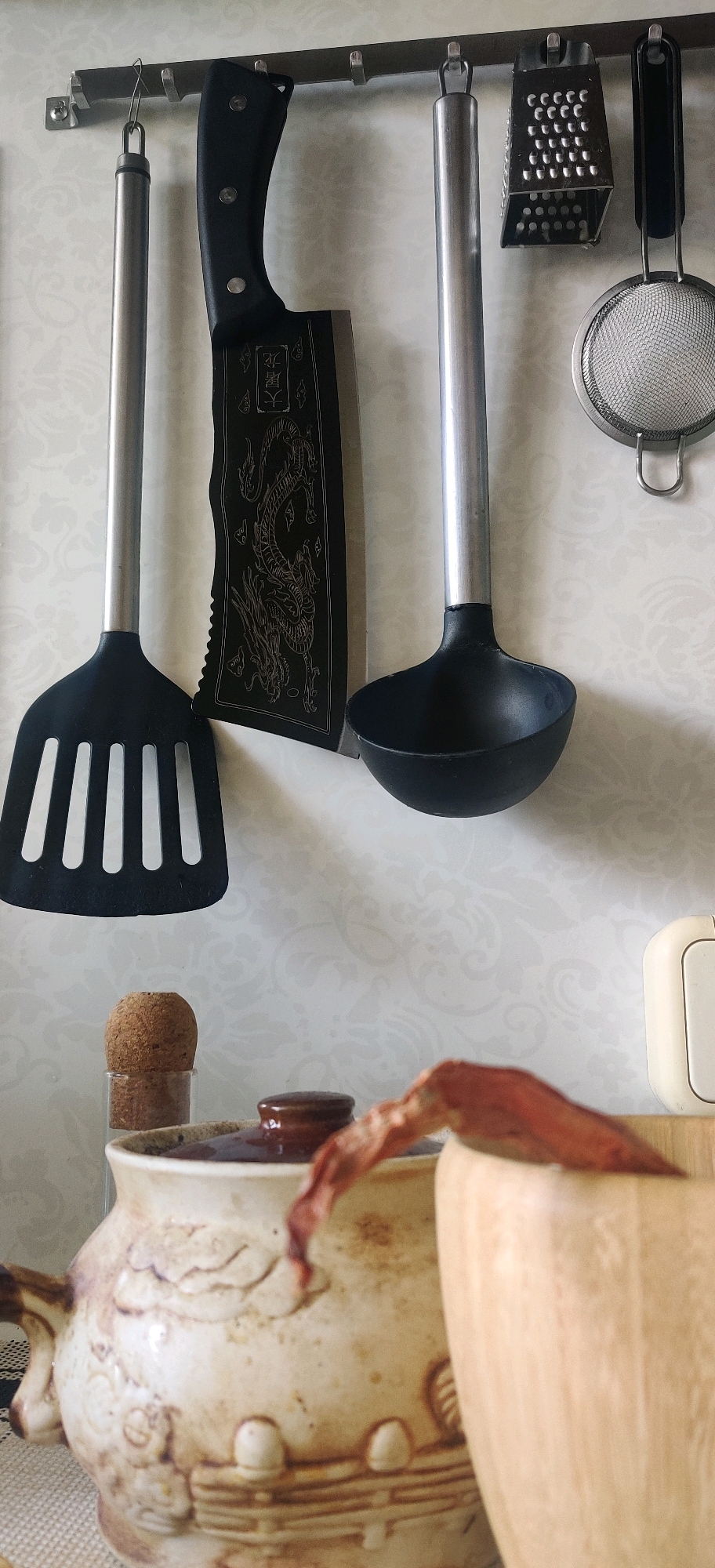 In the city of Hrodna, Belarus, an indoor scene showcases kitchen utensils hanging from a metal rack mounted on a patterned wall. From left to right, a black slotted spatula with a metal handle is suspended. Next to it is a large, black-handled cleaver with intricate dragon engravings on its blade. Further right, a ladle with a black bowl and metal handle hangs, followed by a box grater and a small mesh strainer with a metal handle. Below the hanging utensils, a portion of a ceramic pot with a dark lid is visible, along with a small cork stopper in a clear container. A dried red pepper or similar organic material is draped over the pot and partially obscures a wooden container. The background wall has a subtle, light-colored floral pattern.