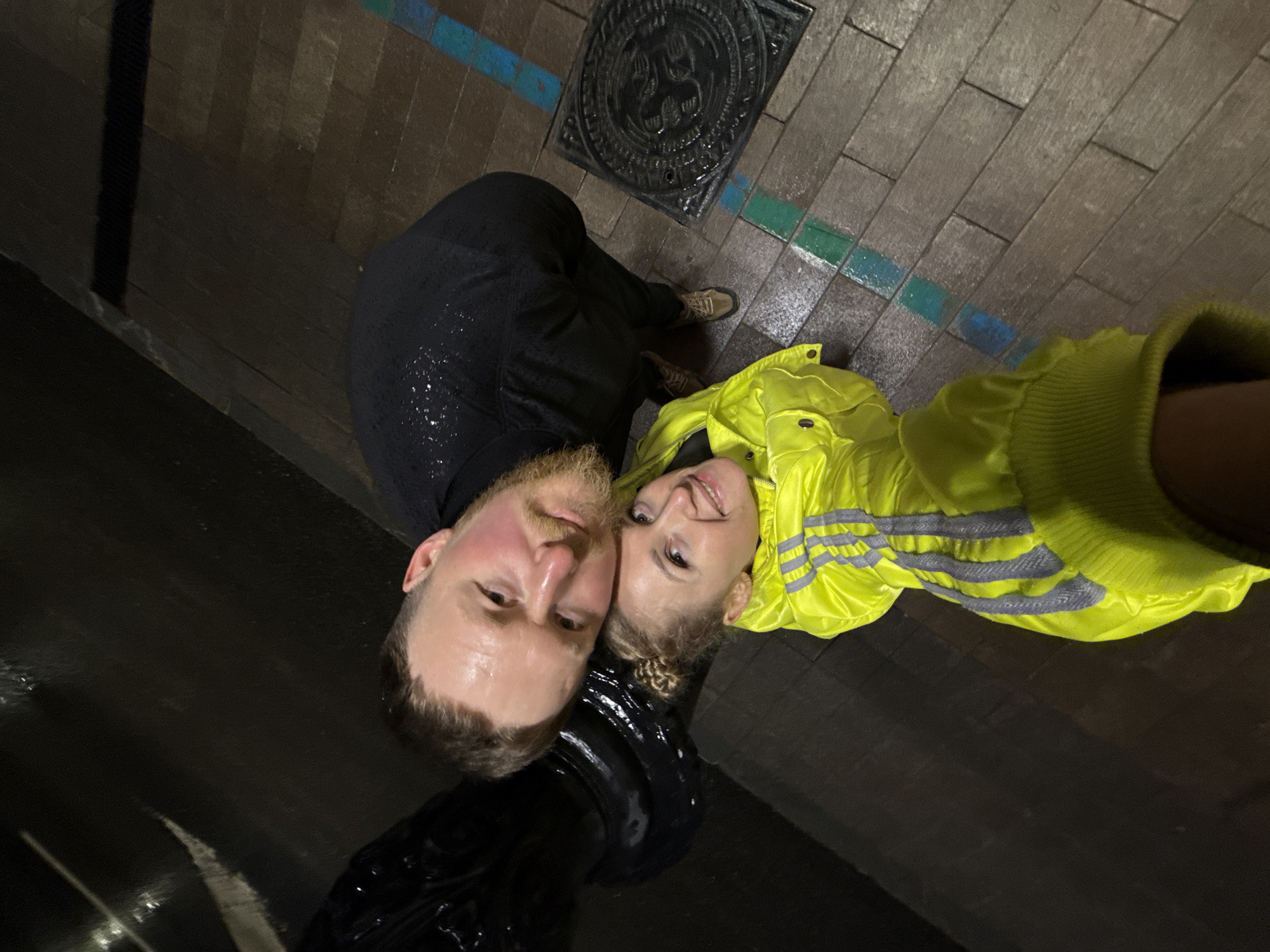 A selfie taken from above shows a Caucasian man and woman posing in an area with wet brickwork, possibly a street in Perm, Russia. The man has a beard and is wearing a dark, wet jacket or sweater. The woman, situated to his right, is wearing a bright yellow rain jacket with reflective stripes. Above them is a cast iron manhole cover set into the brick walkway. The ground reflects some ambient light, indicating wet conditions. There are faint blue and green painted lines running through the brickwork. The overall lighting is low, suggesting it might be dusk or night.