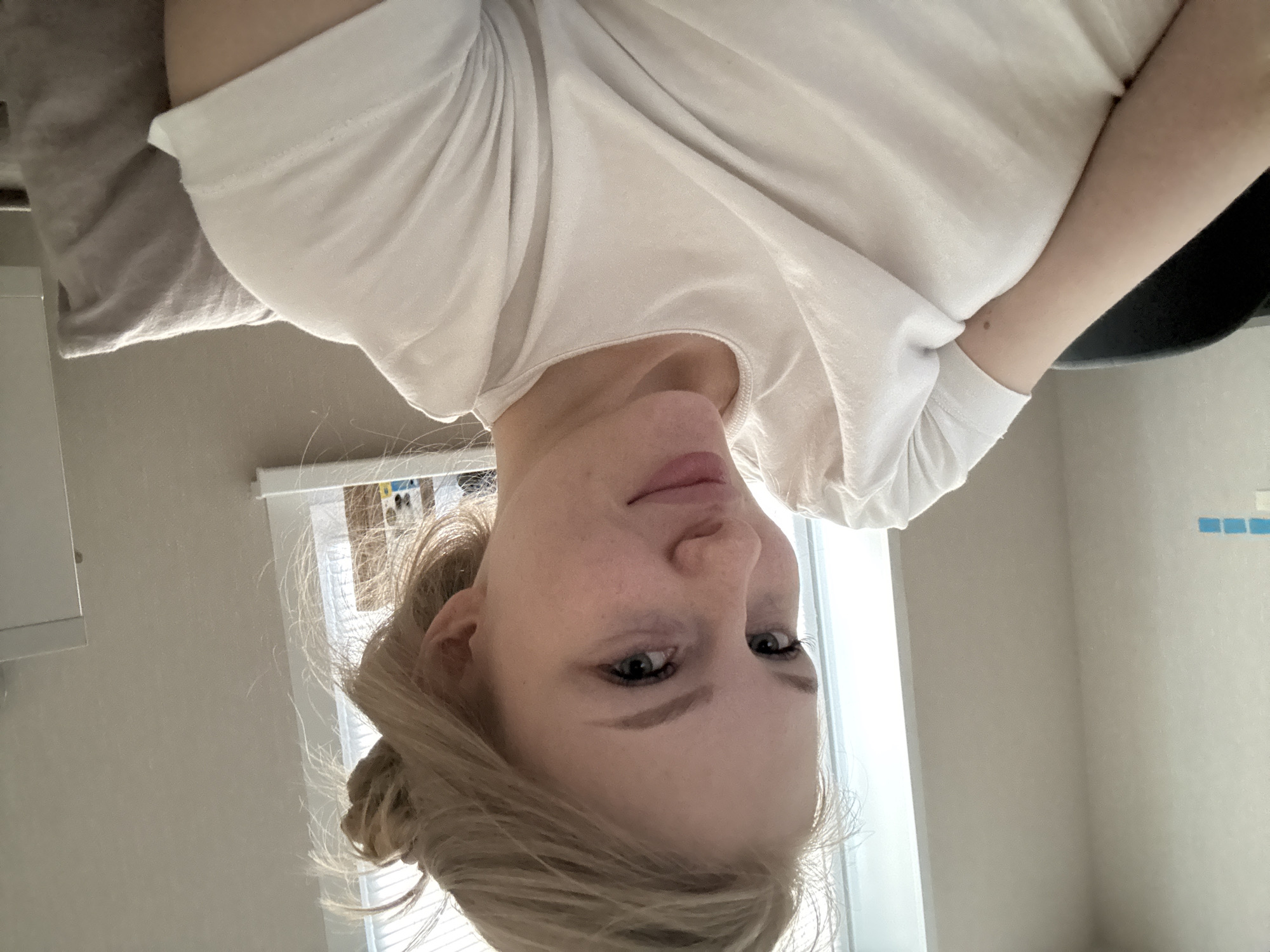 A young woman with blonde hair pulled up in a messy bun is shown from the chest up, looking towards the camera. She is wearing a plain white t-shirt and appears to be indoors. The background is slightly out of focus but suggests a room with a window. The overall lighting is soft and natural. No specific location in the city of Perm, Russia can be determined from the image.